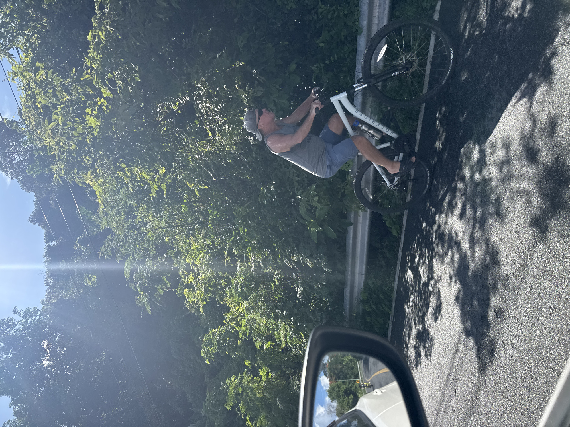 A man rides a white mountain bike on a paved road with green foliage on either side. He is wearing a grey tank top, blue shorts, a grey baseball cap, and sunglasses. A white car is partially visible in the foreground, with its side mirror reflecting the road ahead, showing another vehicle. The sun is shining brightly, casting shadows of trees on the road. The location is identified as Rawai, Thailand, suggesting a tropical or subtropical environment.