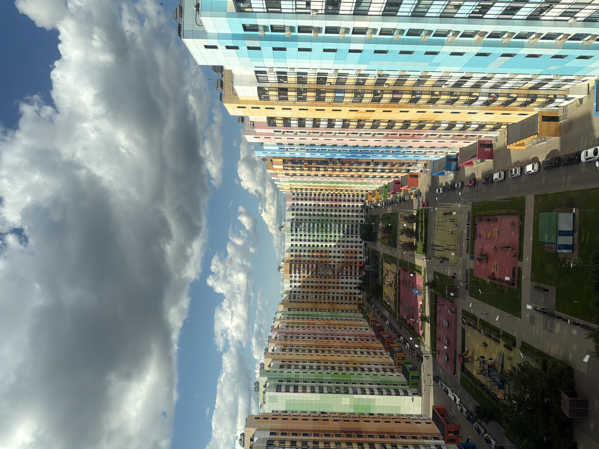 This is a high-angle aerial view of a residential complex in Moscow, Russia. The buildings are tall, modern apartment blocks with brightly colored facades, primarily in shades of orange, green, yellow, pink, blue, and white. The buildings are arranged in a U-shape, enclosing a large courtyard area. Within the courtyard are multiple playgrounds with colorful equipment, paved walkways, green spaces with trees and benches, and parking areas. Several cars are parked along the street bordering the complex and within the courtyards. The sky is partly cloudy with dramatic, grey clouds interspersed with patches of blue sky. The overall impression is of a vibrant and newly developed urban area.