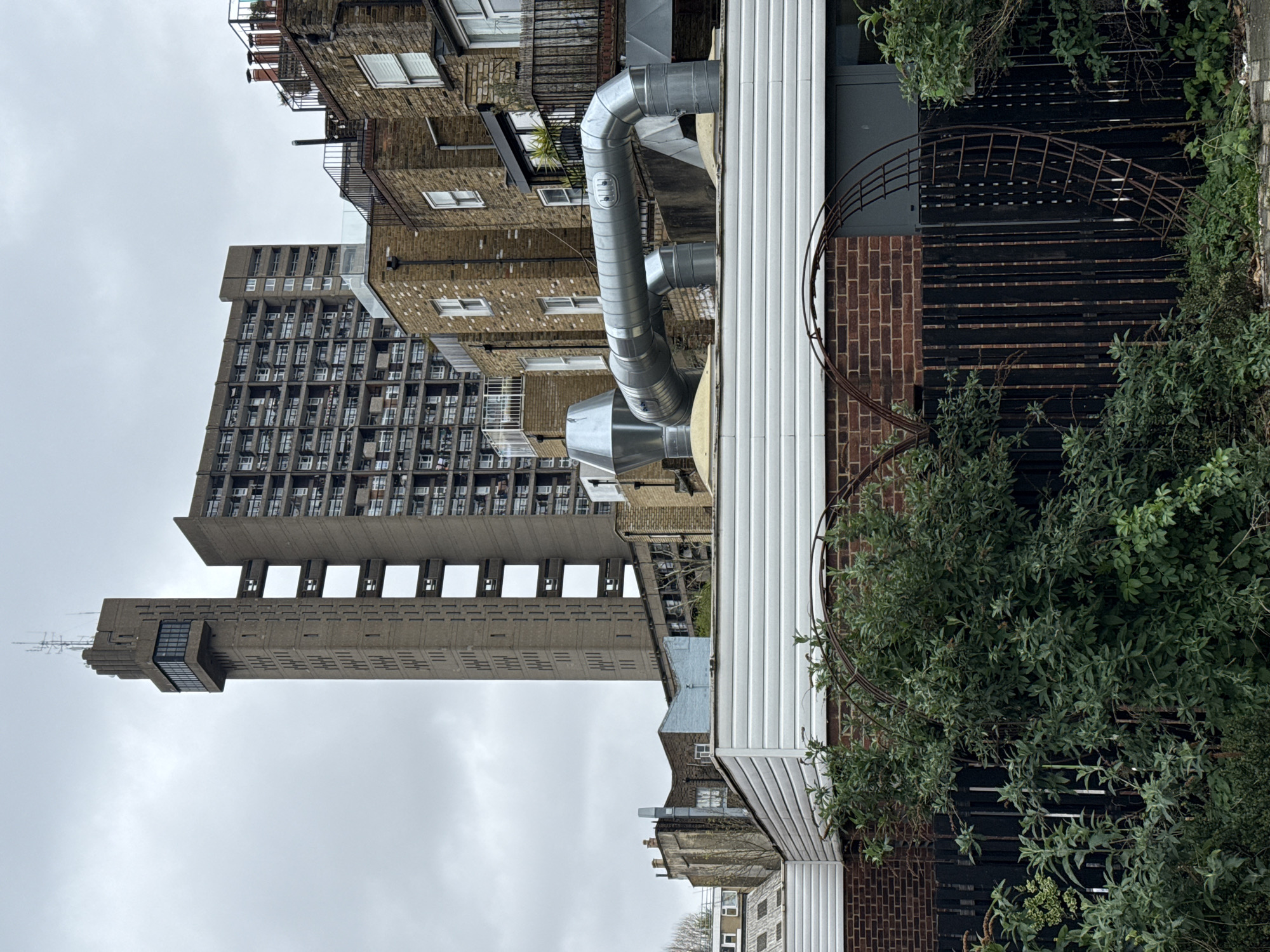 The image depicts a scene in London, United Kingdom, showcasing a juxtaposition of architectural styles. A prominent, high-rise brutalist building with visible concrete structure is visible on the left. Adjacent to this is a more traditional brick building with balconies and windows, suggesting a residential function. A series of industrial-looking, galvanized metal ducts snake across the roof of a building. In the foreground, a wooden fence with brick accents and green shrubbery provides a sense of enclosure. The sky appears overcast.