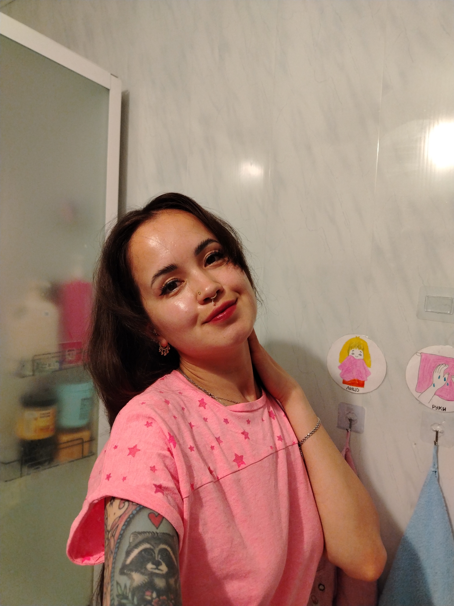 The image shows a young woman in a pink t-shirt with stars, standing in a bathroom. She has a nose ring and is wearing earrings. Her left arm is tattooed with a raccoon and other designs. In the background, there are shelves with toiletries and two circular drawings on the wall, one depicting a girl and another of hands washing. The overall environment suggests a domestic setting. The city of Noviy is not depicted or referenced in the image.