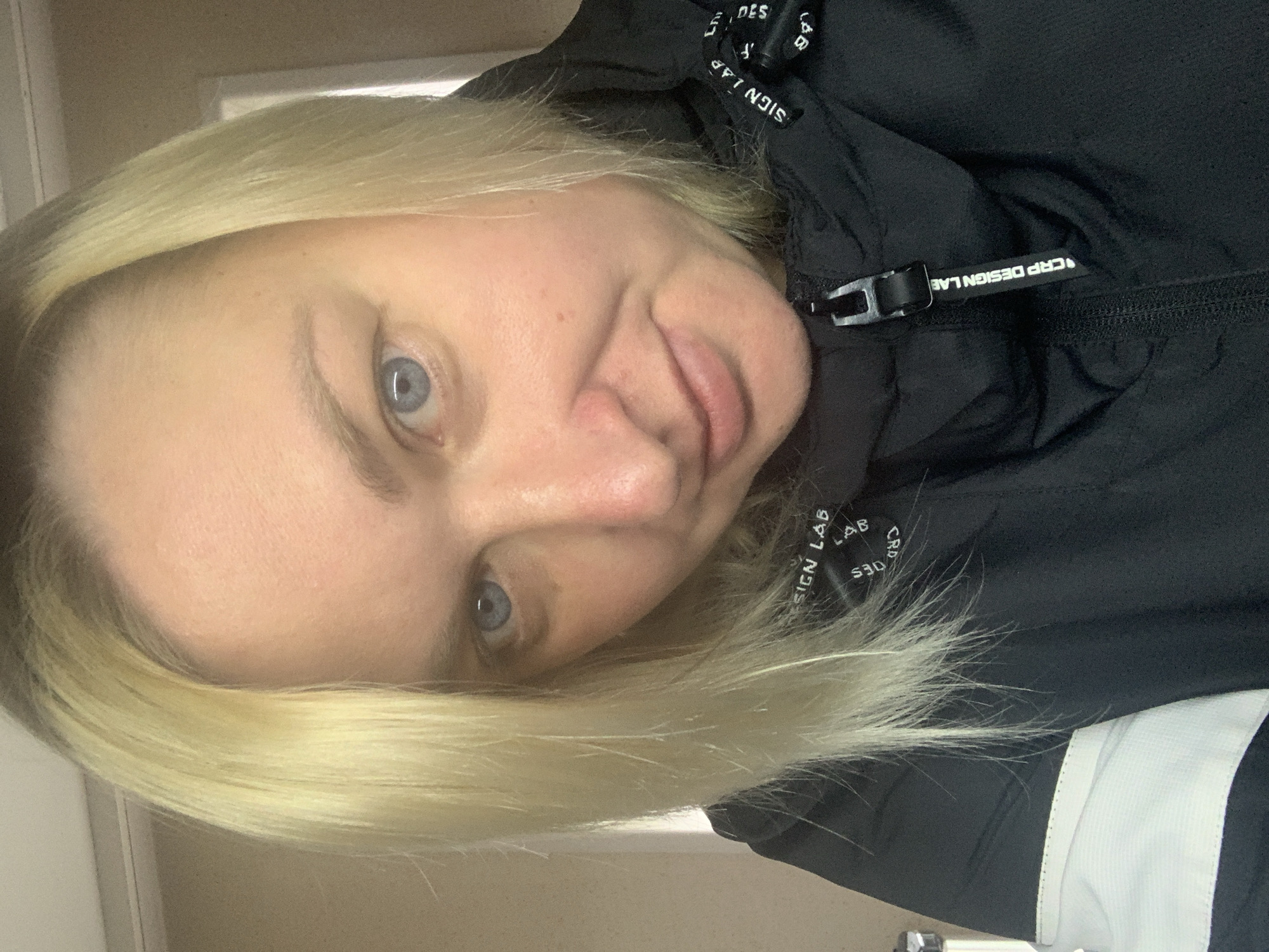 A close-up, tilted portrait features a person with blonde hair and light blue eyes, situated in Chaikovsky, Russia. The individual looks directly at the camera with a neutral to slightly relaxed expression, her lips slightly parted. Her blonde hair is styled to frame her face, with some strands falling across her forehead.
She is wearing a black jacket, likely a hooded type, as indicated by visible straps and details around the neck/hood area. These black straps are adorned with white text, clearly showing phrases such as "CRP DESIGN LAB", "LAB", "DESIGN", and "CRP" repeated and oriented at different angles. A black zipper runs down the front of the jacket, and a black plastic buckle or zipper pull, also imprinted with "CRP DESIGN LAB" in white, is visible near the top of the zipper. A segment of a white panel or strip on the jacket's right side adds a contrasting detail.
The background is a simple, light-colored interior wall or door frame, suggesting the photo was taken indoors. The lighting is even and soft, providing no specific clues about the time of day or outdoor weather conditions. The image captures a casual and direct gaze.