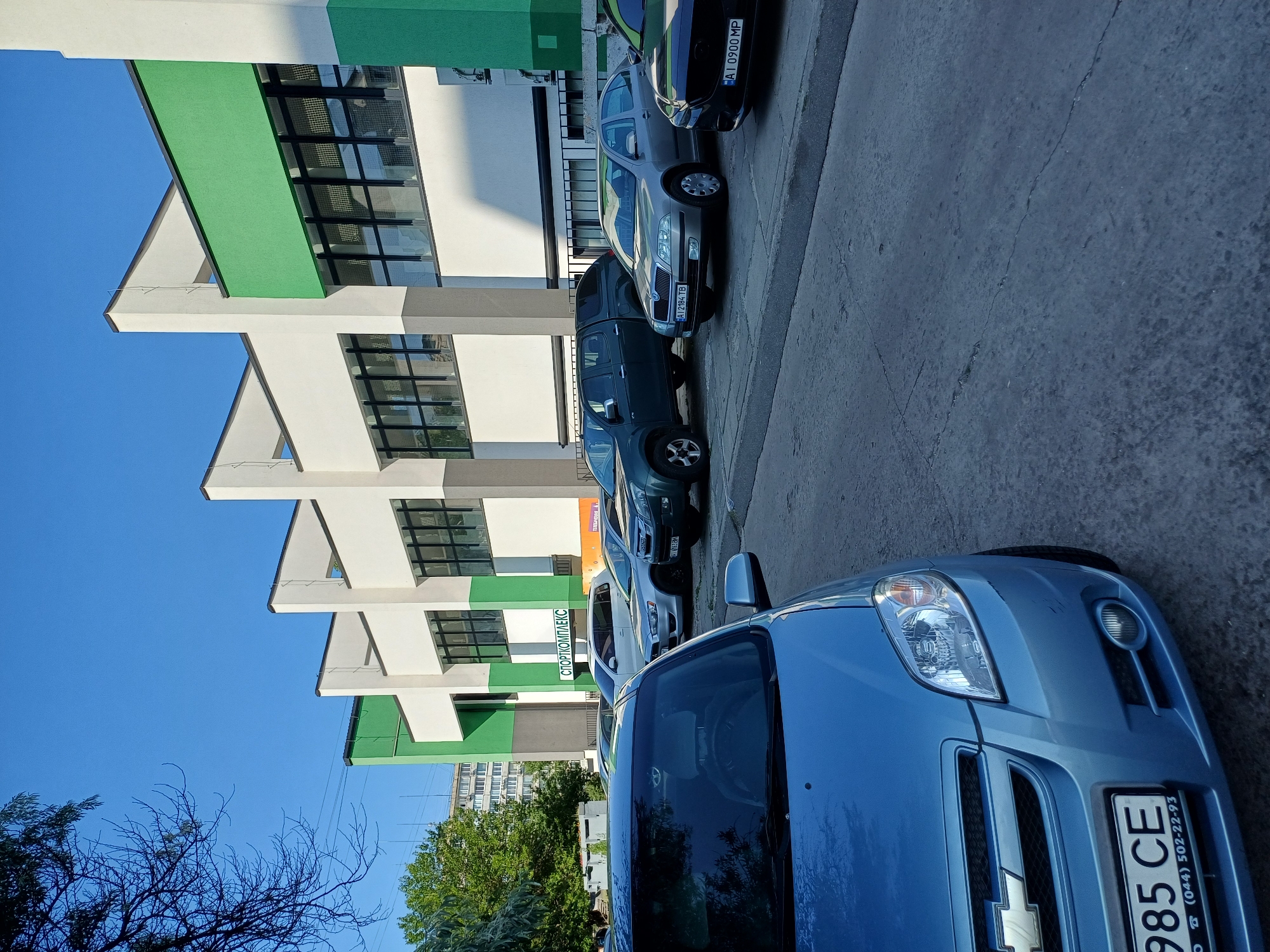 In Ukrainka, Ukraine, a blue Chevrolet Aveo is parked in the foreground facing left. The car has a license plate reading "985 CE" and phone number "(044) 502-22-93". Behind it, several other cars are parked, including a silver Skoda Octavia with license plate "AI 2184 TB", a dark green Isuzu D-Max pickup truck with a canopy, and a dark sedan with license plate "AI 0900 MP". The cars are parked on a concrete surface in front of a modern building with a white facade and green accents. A sign above the entrance reads "СПОРТКОМПЛЕКС" (SPORTS COMPLEX). The sky is clear and blue. Trees are visible to the left of the building.