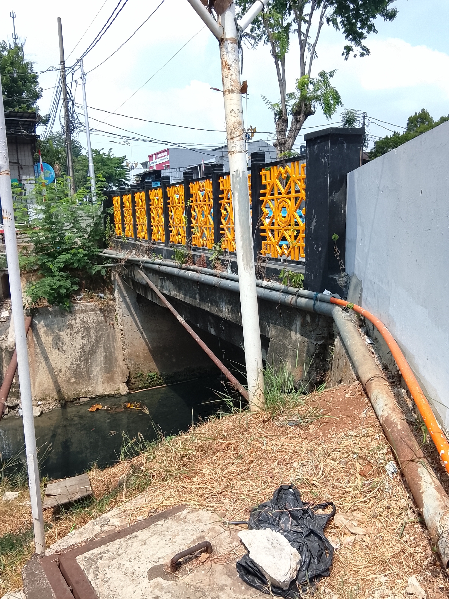 The image shows a narrow canal with murky water flowing under a concrete bridge. The bridge is supported by concrete walls and has rusty pipes running along its side. A bright orange pipe is also visible on the right side of the image, connected to the bridge. The area surrounding the canal is overgrown with dry grass and some green bushes. A black plastic bag with some debris is on the ground near the canal. In the background, a black fence with an intricate yellow pattern runs along the top of the canal. Beyond the fence, there are trees, utility poles with numerous wires, and buildings, including one with a red facade. The sky is partly cloudy. The scene appears to be in an urban or semi-urban environment in East Jakarta, Indonesia.
