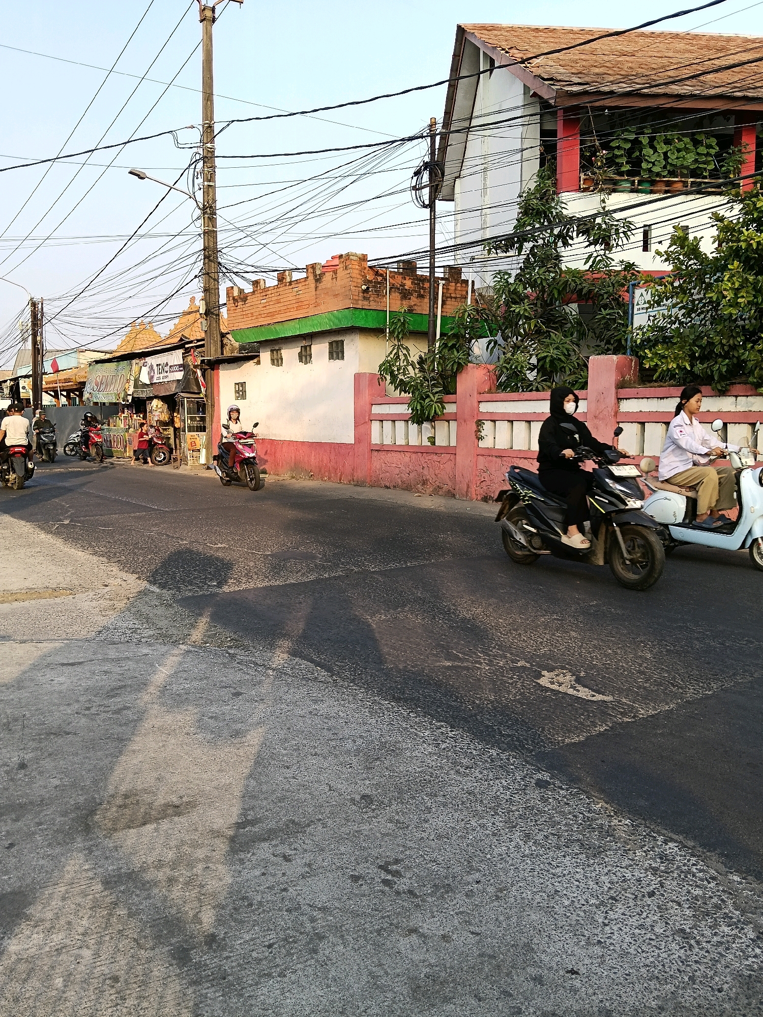 The image depicts a street scene in Belimbing, Indonesia. The foreground shows a road with a rough, textured surface, reflecting sunlight and casting shadows. Several individuals are riding motorcycles. On the right side of the road, two women are on motorcycles; one is wearing a black hijab and a black outfit, and the other is wearing a light-colored shirt and tan pants. Behind them, on the left side of the road, several other motorcycles are visible, some with multiple riders. Businesses with storefronts line the right side of the street, featuring signs and awnings. The buildings have red-tiled roofs and pink and white facades. A prominent utility pole with numerous tangled wires is situated on the left side of the street. Green foliage is visible on some of the buildings, and trees are scattered throughout the scene. The sky is clear and light blue, suggesting it is daytime.