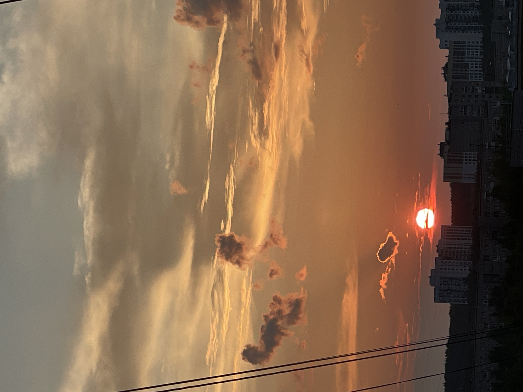 The image shows a sunset over a city skyline, likely Izhevsk, Russia, given the architectural style of the buildings which appear to be Soviet-era apartment blocks. The sky is filled with dramatic clouds, painted in hues of orange, yellow, and pink by the setting sun. A bright, circular sun is partially obscured by a cloud formation near the horizon. In the foreground, silhouetted against the vibrant sky, are several tall, multi-story buildings, characteristic of residential areas. Darker, lower buildings and trees are visible at the base of the frame. There are also dark, vertical lines on the left side of the image, possibly representing power lines or wires. The overall impression is one of an urban landscape bathed in the warm, atmospheric light of a sunset. There are no visible people in the image.