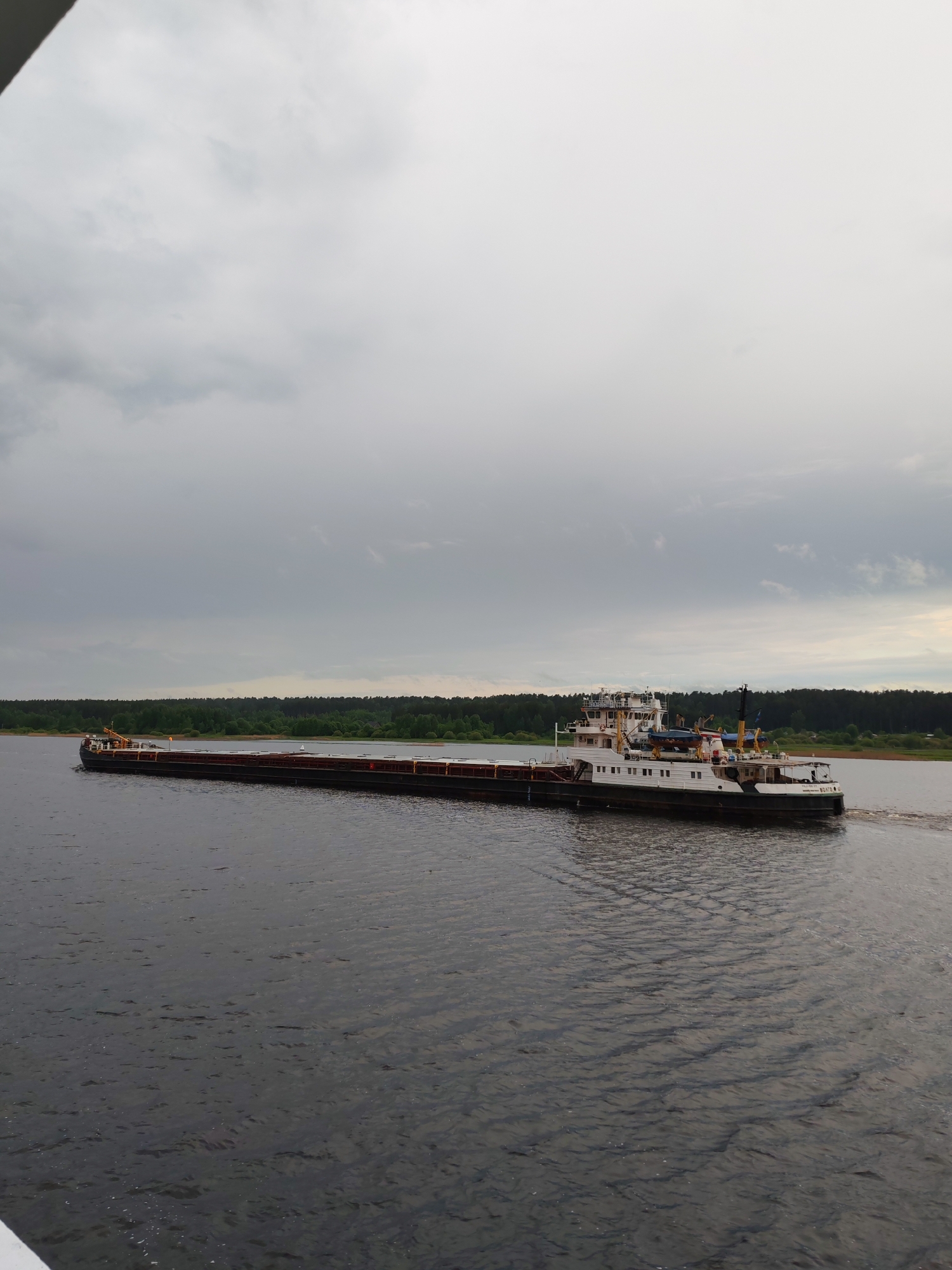 An overcast sky hangs over a wide body of water, likely the Volga River, in Myshkin, Russia. A long, dark barge is being pushed by a tugboat with a white superstructure. The tugboat is positioned at the rear of the barge. Dense green trees line the far bank of the river.