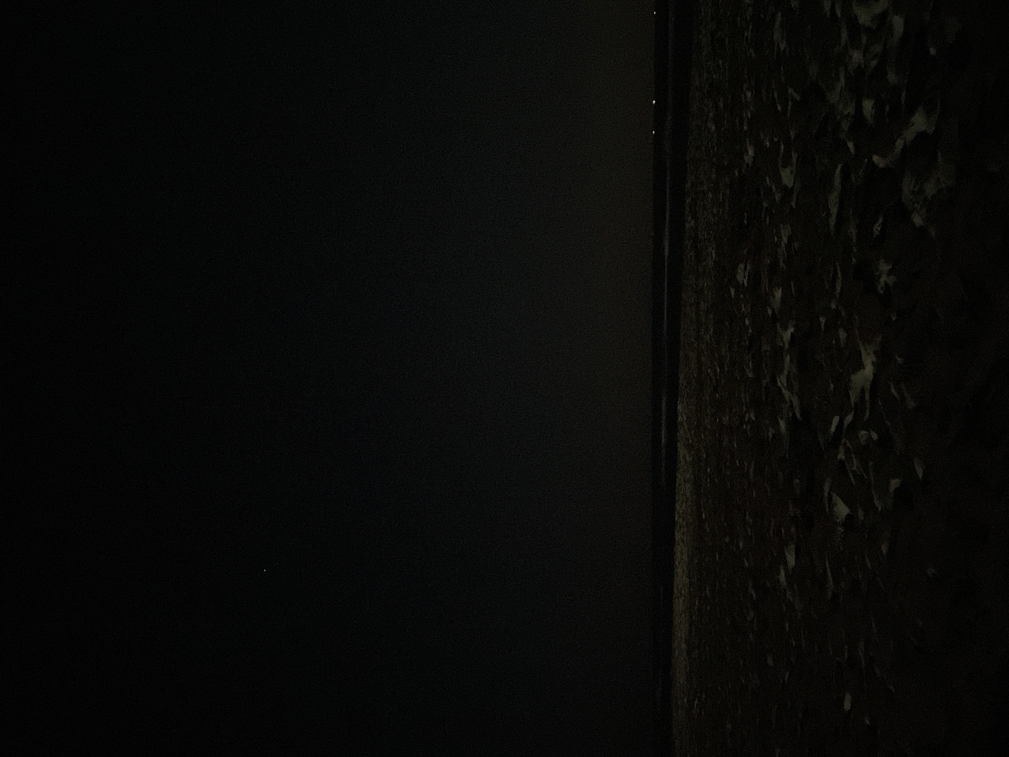 The image depicts a dark, expansive scene, likely taken at night. The foreground is dominated by what appears to be a sandy or uneven surface, with patches of lighter material scattered across it, possibly snow or ice. This textured area occupies the majority of the lower half of the frame.
Above the textured foreground, a dark horizon line separates it from a vast expanse of blackness that fills the upper portion of the image. This upper area is largely featureless, suggesting a clear night sky. There are a few very small, faint points of light visible in the upper portion of the frame, one slightly larger and brighter in the upper left quadrant, and two smaller, dimmer lights near the horizon on the right. These could be stars, distant lights, or possibly aircraft.
The overall impression is one of darkness and solitude. The lack of distinct features and the deep shadows make it difficult to ascertain the exact nature of the environment or the presence of any specific objects beyond the surface texture in the foreground. The scene is devoid of any visible human presence or activity. The location is identified as Unknown, Greece, but there are no visual cues within the image to support or refute this.