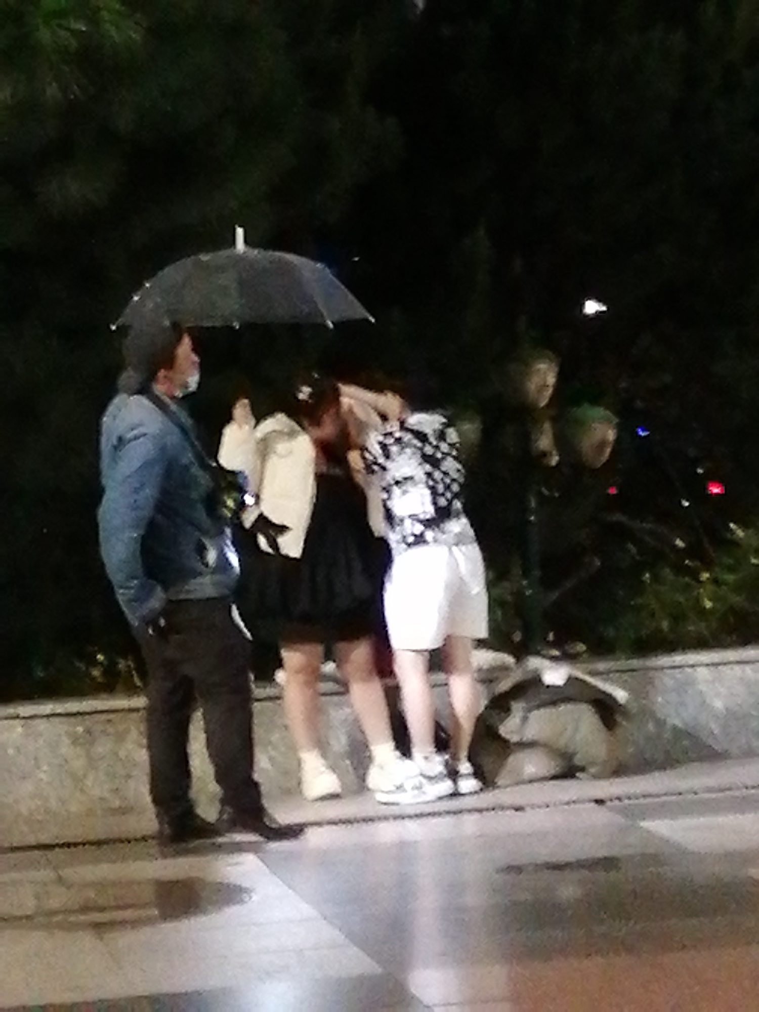 The image depicts a dimly lit outdoor scene, likely at night, with a group of individuals gathered under a black umbrella. The location is identified as Da Lat, Vietnam, with a wet paved area in the foreground suggesting recent rain.
In the center-left, a man wearing a blue denim jacket and dark pants stands facing away from the viewer, holding the umbrella. To his right, a person in a white top and dark skirt is partially obscured by the umbrella. Further right, another individual is dressed in a patterned short-sleeved shirt and white shorts. Behind them, indistinct figures are visible in the background, appearing to be in a lush, dark environment with hints of foliage and possibly some colorful lights. A large, light-colored bag or bundle is situated on the ground to the right of the group. The overall impression is a candid snapshot of people seeking shelter from the elements or simply conversing outdoors.