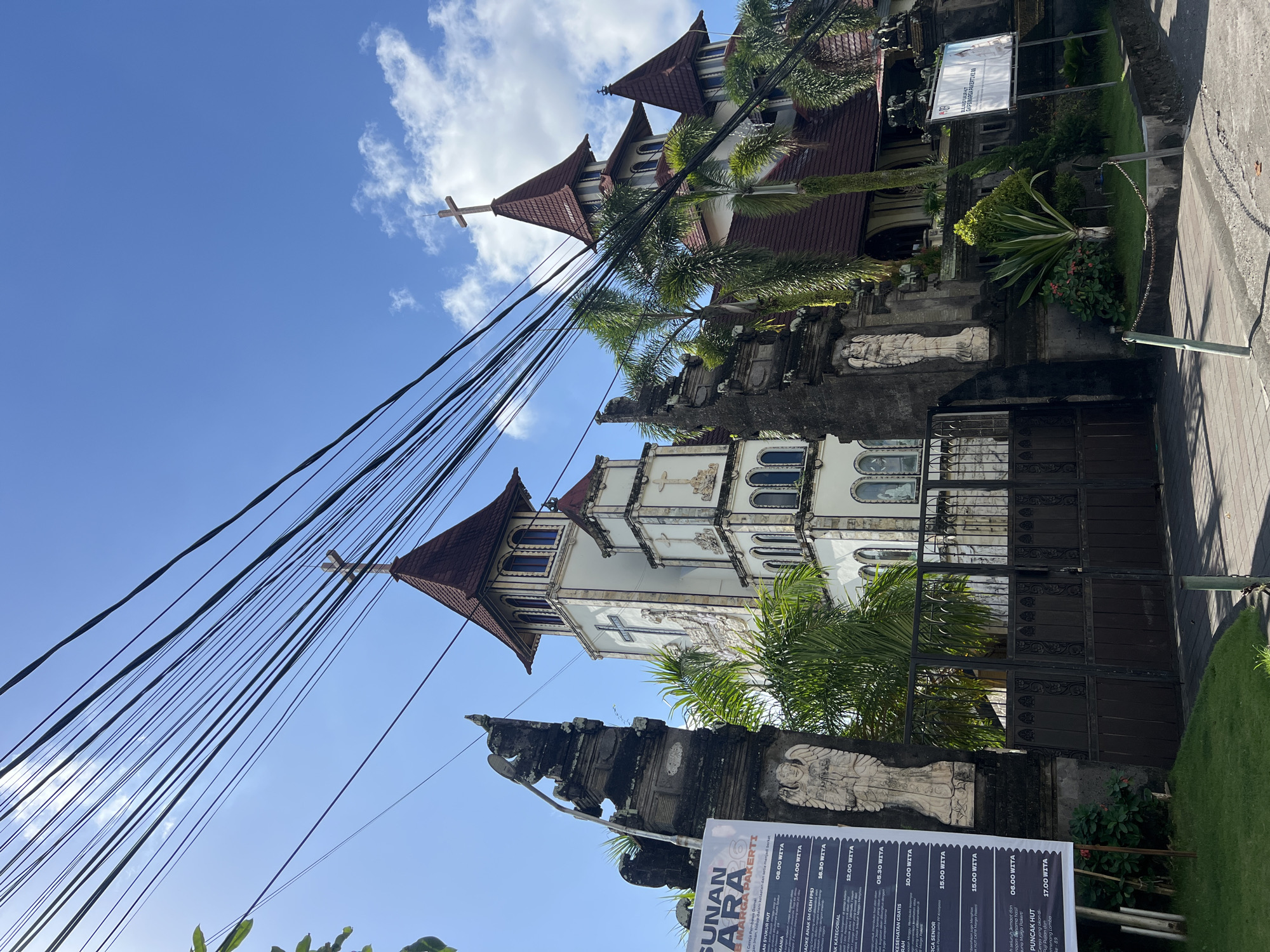 The image shows the exterior of the Gereja Katolik Maria Bunda Karmel church in North Kuta, Indonesia. The church features a tall, white tower with a red, conical roof, topped with a cross. Adjacent to the tower are other sections of the church with similar architectural elements. The entrance to the church grounds is marked by ornate stone gates with statues. A dark metal fence encloses the gate. Overhead, a dense network of black electrical wires crisscrosses the bright blue sky, which is partially filled with white clouds. Palm trees and other greenery are visible on the church grounds. A white sign with text and an image is mounted on the wall to the right of the entrance. Another poster is displayed on a stand to the left of the gate. The scene is illuminated by daylight.