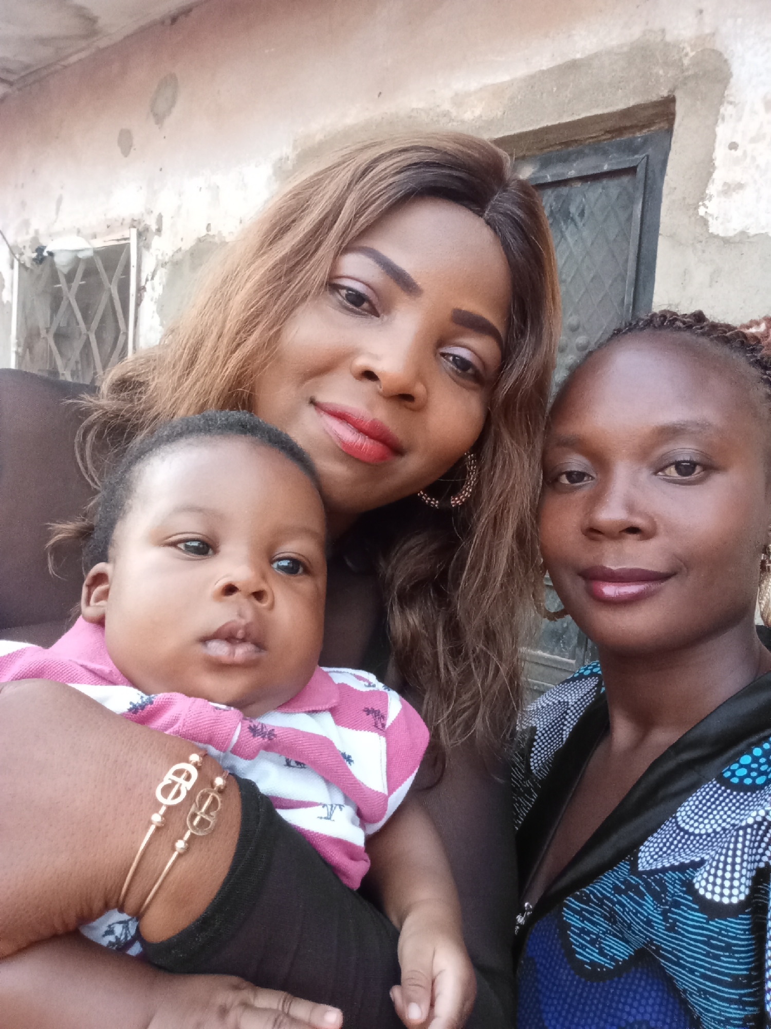 Three individuals are depicted in the photograph. The composition is a close-up shot, seemingly taken by someone holding the camera at arm's length.
In the foreground, a baby is held by an individual with a dark-skinned complexion. The baby is wearing a pink and white striped shirt. The arm holding the baby has a gold bracelet.
Two adult women are visible behind the baby. The woman on the left has brown hair and red lipstick. The woman on the right has braids and is wearing a patterned blue and black top.
The background consists of a building with a weathered exterior. The building has a window with a metal grill and a dark colored door. The location is presumed to be Sabon Tasha, Nigeria.
