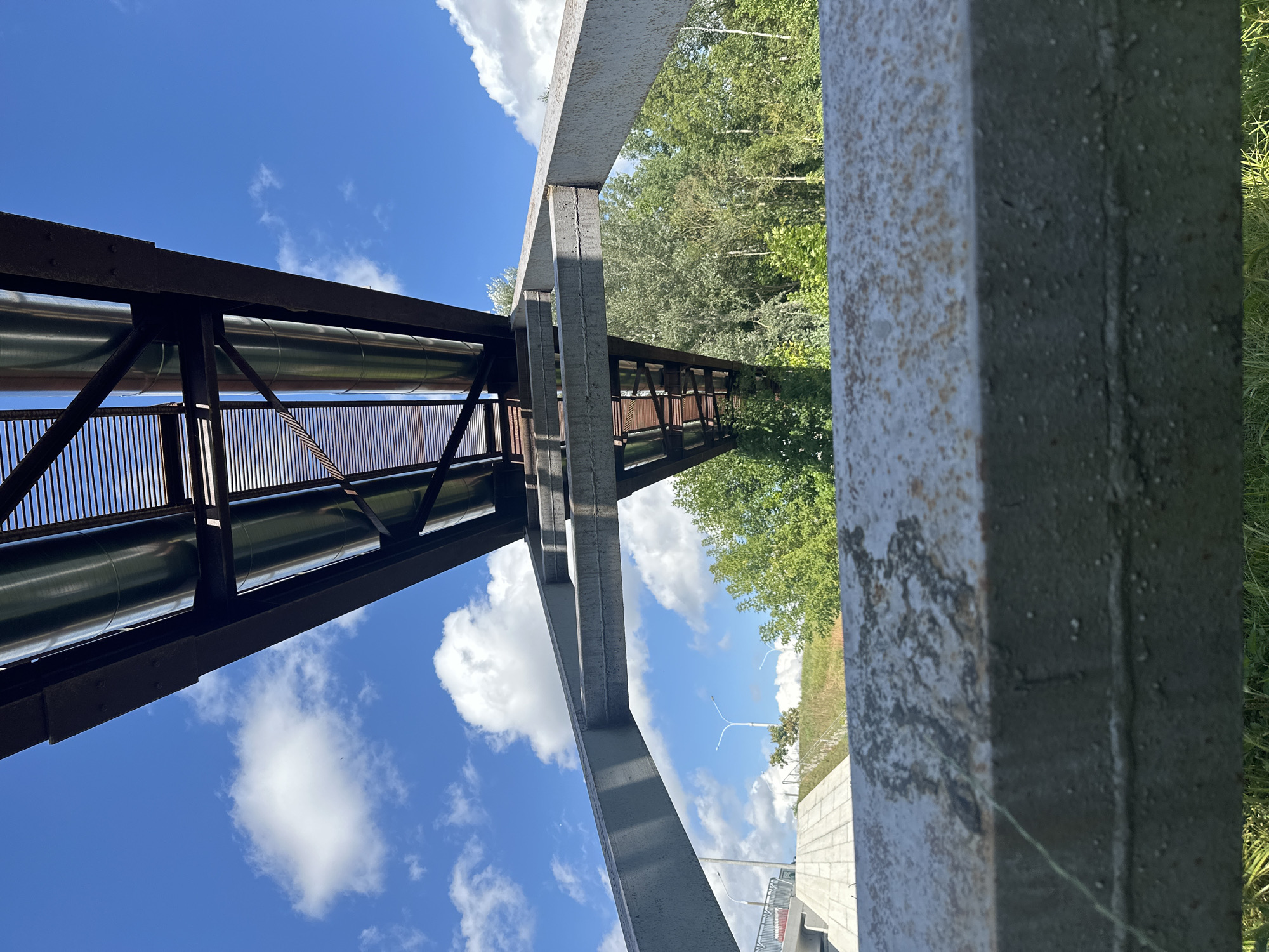 The image shows an underside view of an industrial pipeline structure against a bright blue sky with scattered white clouds. Two large, silver-colored pipes are visible, supported by a dark metal framework that includes horizontal bars and vertical beams. Below the pipes, a lighter gray, angled beam forms part of the larger structure. The foreground features a weathered, gray metal railing or barrier, slightly out of focus, with visible signs of rust and texture. In the background, beyond the pipeline structure, a tree line of lush green foliage is present. To the left, a concrete ramp or pathway slopes upwards, leading towards streetlights and what appears to be a bridge or overpass. The scene is outdoors during the daytime, under sunny conditions, with shadows cast by the pipeline structure. The location is identified as Minsk, Belarus.