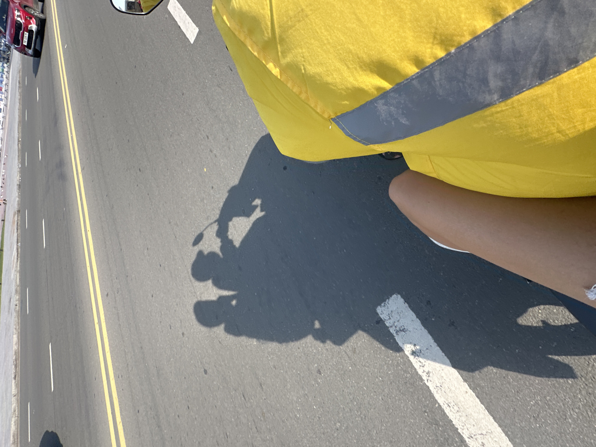 The image captures a top-down, slightly forward perspective from a person riding a motorcycle or scooter on a sunny day. The rider's bare right leg is visible in the lower right, along with a portion of a bright yellow garment, possibly a rain poncho or jacket, which features a grey reflective strip.
The asphalt road is the dominant feature, marked with a prominent double yellow line indicating no passing, and white solid and dashed lines along the side. A large, distinct shadow of the motorcycle and rider is cast across the road, clearly showing the handlebars, a mirror, and the outline of a person.
On the left side of the road, a red car, possibly a Kia Seltos, is parked next to a sidewalk. The car's license plate, "50G-200.79", is visible. Further down the sidewalk, several small birds, likely pigeons, are scattered. A rearview mirror is partially visible in the top left, reflecting a distorted view of the surroundings.
The strong shadows and bright illumination indicate clear, sunny weather, likely around midday or early afternoon. The scene takes place on a road in Nha Trang Ward, Vietnam.