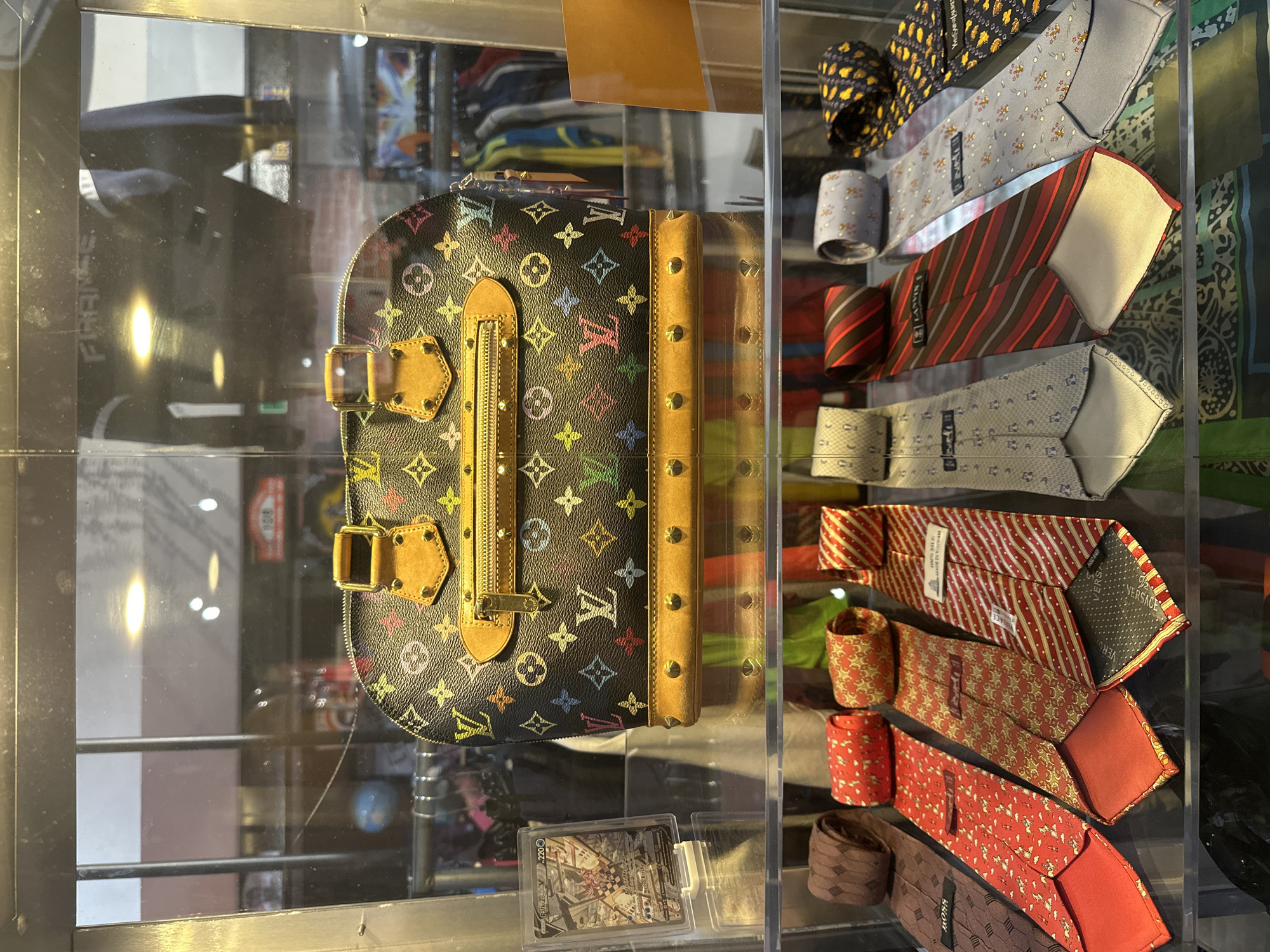 This image depicts a display case filled with various luxury items. At the top, a black Louis Vuitton Speedy bag with the iconic multicolor monogram pattern is prominently displayed. The bag features gold-toned hardware and tan leather trim. To the left of the bag, a trading card in a clear plastic case is visible. Below the bag, several rolled-up ties are arranged on a shelf. The ties showcase a variety of colors and patterns, including stripes, small prints, and a brown patterned tie. The display case appears to be situated in a retail environment, with racks of clothing visible in the background. There are no people directly visible in the image. The overall impression is that of a curated collection of fashion accessories, likely in a boutique or vintage store. The image does not contain any information or context related to the city of Dieppe, France.
