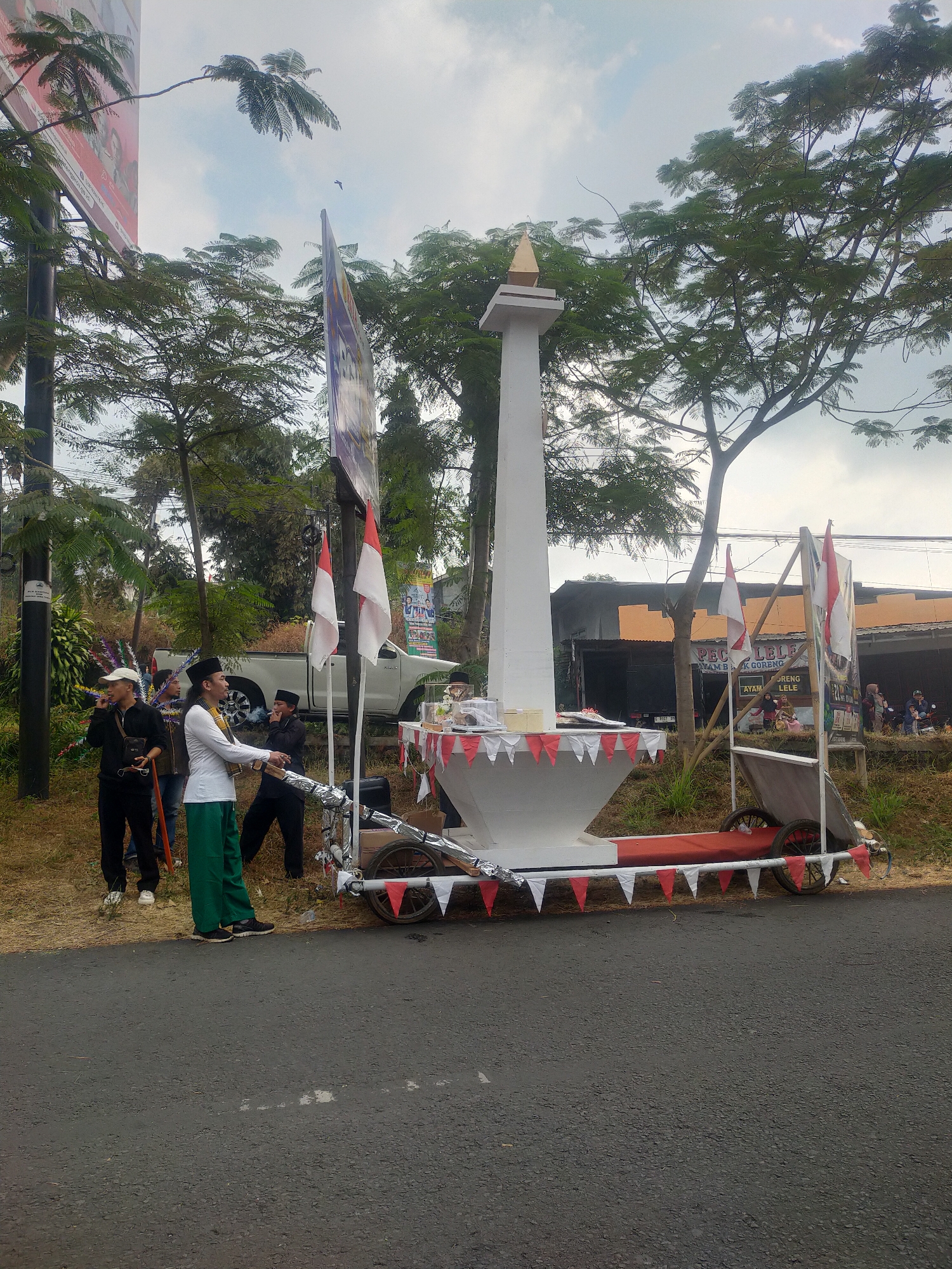 In Tuwel, Indonesia, a temporary monument replica, resembling a white obelisk with a golden finial, stands on a wheeled platform decorated with red and white triangular flags. The monument is positioned on the side of a road, with a white pickup truck parked behind it. A group of people are gathered around the monument, some in traditional attire. One man, wearing a white long-sleeved shirt and green pants, is holding a microphone and appears to be speaking or singing. Another man in a white cap and black jacket is also holding a microphone. The environment features lush green trees, a cloudy sky, and a roadside building with a sign that reads "PEC LELE." The scene suggests a local celebration or event.