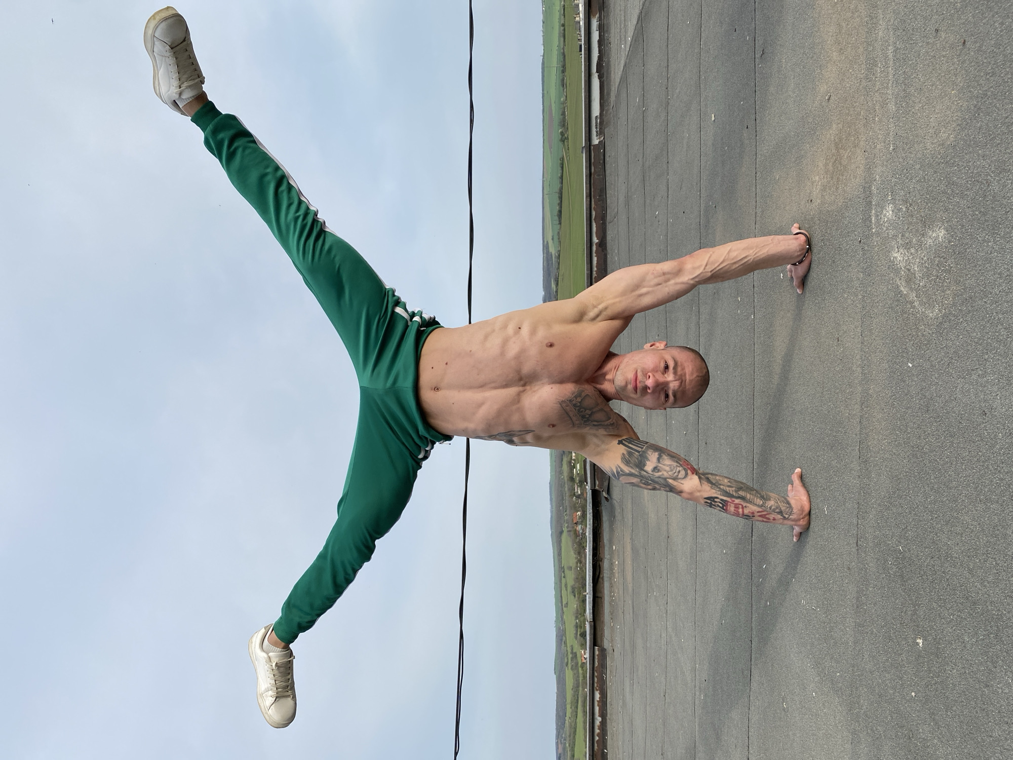 A muscular man with tattoos is performing a handstand split outdoors. He is wearing green sweatpants and white sneakers. He is balancing on his hands on a grey surface, possibly a rooftop. Beyond the rooftop, a landscape with green fields and trees is visible. The sky is overcast. The location is in Pleven, Bulgaria, based on the provided information.
