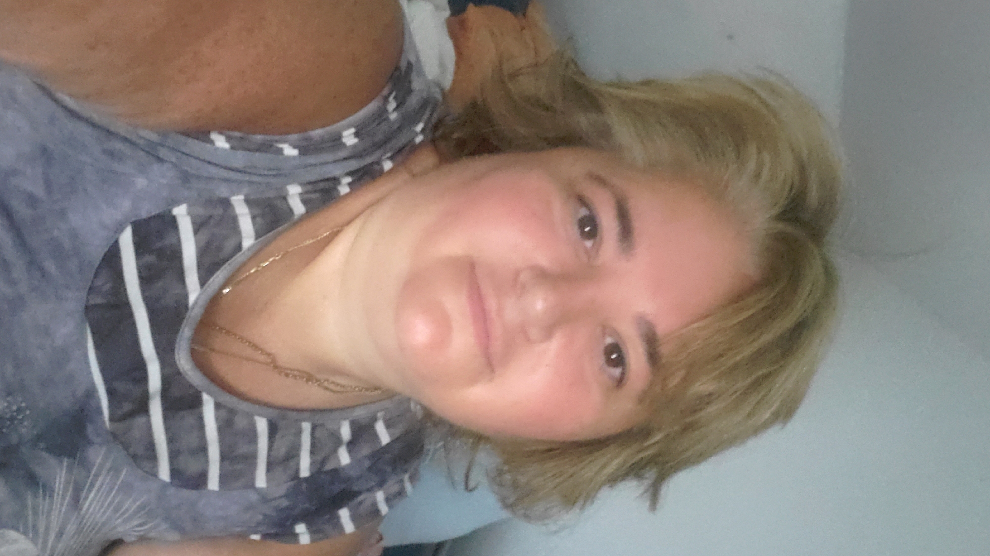 A woman with blonde hair is pictured in a close-up shot, facing the camera. She is wearing a grey and white striped t-shirt. The background appears to be an indoor setting with a light-colored wall and some blurred objects to the left. The woman has a necklace on. No location in Sasbach, Germany is discernible from the image.