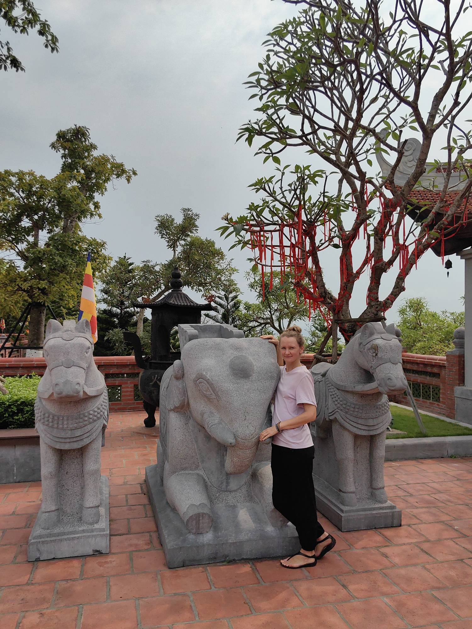 A Caucasian woman stands alongside a gray stone elephant sculpture, which is flanked by two gray stone horse sculptures, on a red brick patio in Hong Gai Ward, Vietnam. The woman wears a light pink short-sleeved shirt, black pants, and dark sandals. A bare tree with red ribbons tied to its branches stands behind her and the elephant sculpture. In the background are a black incense burner, a flagpole with a yellow-red striped flag, various other trees, and a red-tiled roof with gray trim. The sky is overcast.