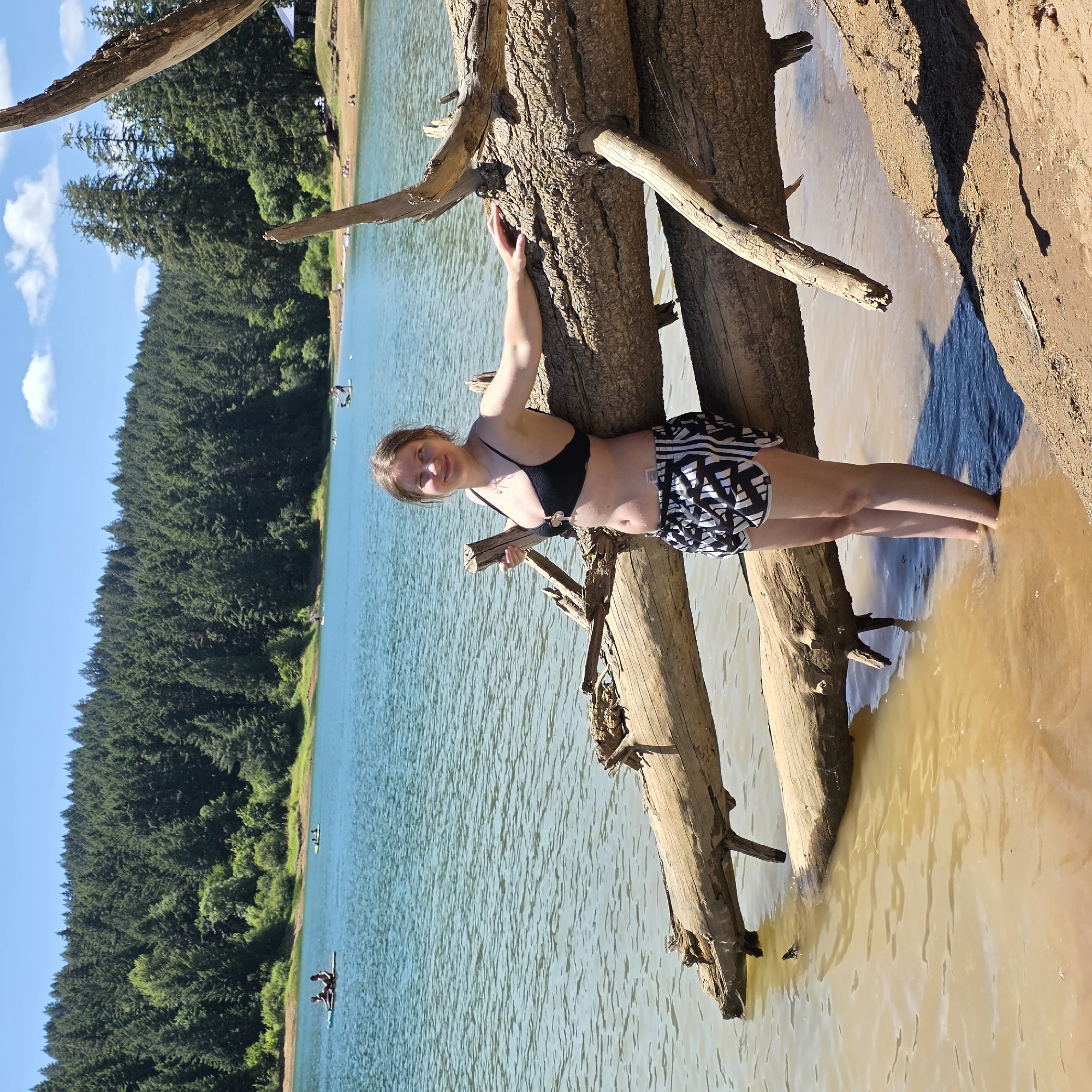 In Cherry Grove, Oregon, a young woman stands at the edge of a body of water, partially submerged, with her feet in the shallows. She is leaning against a large, fallen tree trunk that lies horizontally across the water and the shore. The woman is wearing a black bikini top and black and white patterned shorts. Behind her, the lake stretches out, bordered by a dense forest of evergreen trees that climb a hillside. The sky is blue with scattered white clouds. In the distance on the water, several people are visible, some on paddleboards and others in small boats. The shore is sandy with some exposed tree roots and debris. The overall scene depicts a sunny day at a natural lake with recreational activities.
