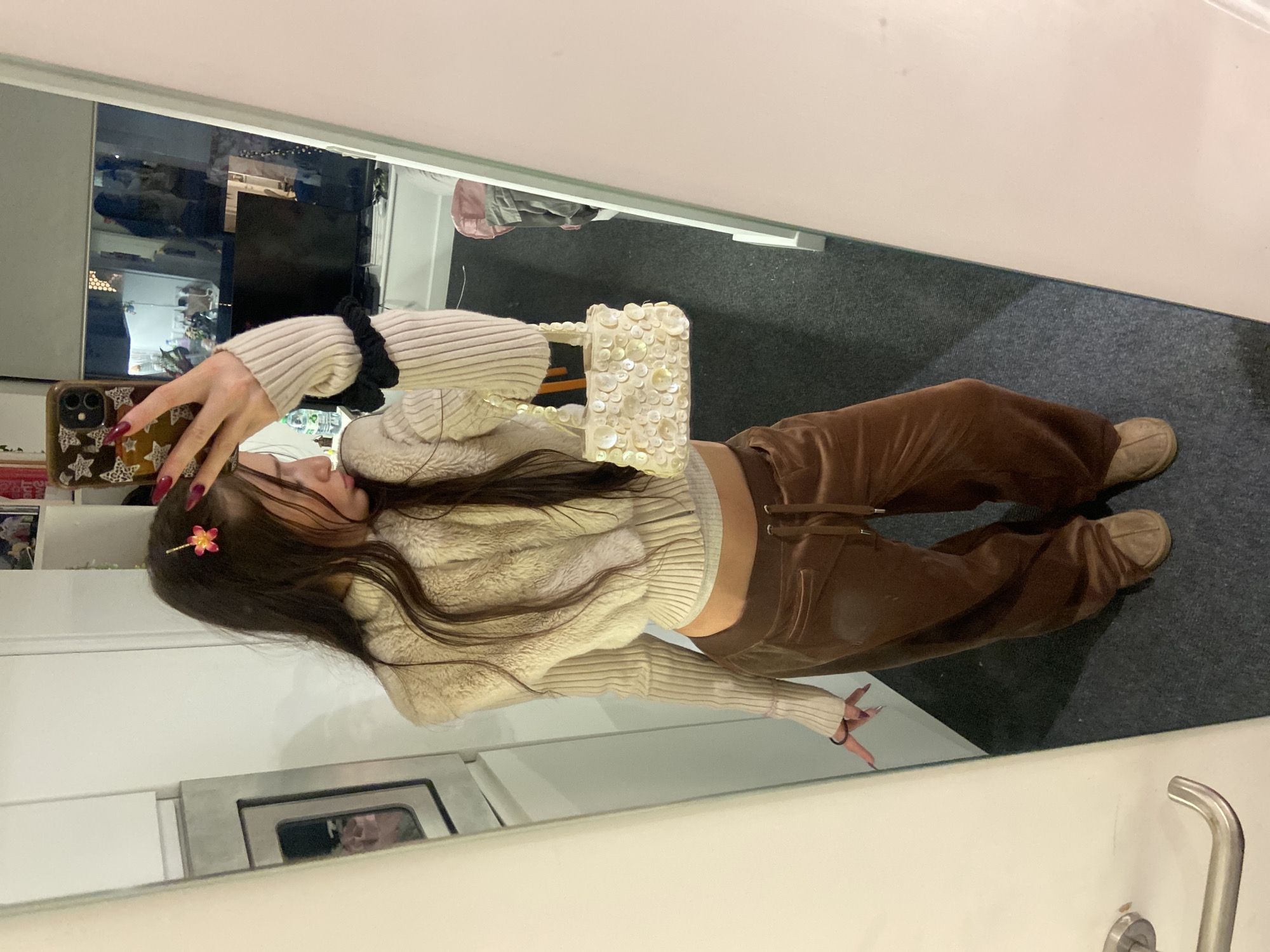 A young woman is taking a selfie in front of a mirror. She is wearing a cream-colored fuzzy sweater over a ribbed top, brown velvet pants, and beige shoes. She is holding a white beaded purse. A flower hair clip is visible in her hair. The background appears to be a room with a white cabinet and a glimpse of another room with furniture. The location is the United Kingdom, and the person is taking the image in a mirror that shows a London street.
