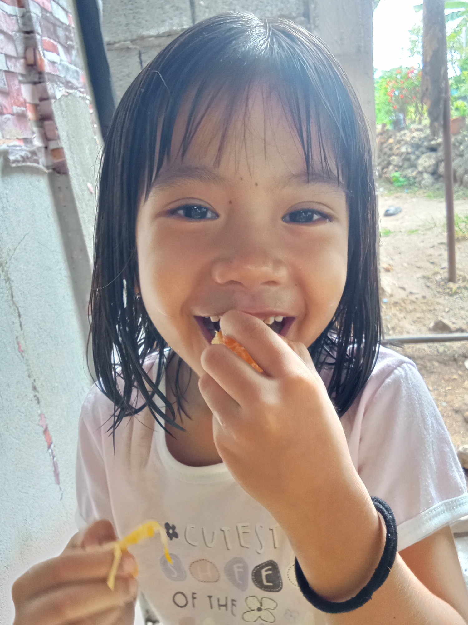 A young girl with dark hair is smiling and eating a piece of orange fruit. She is wearing a light pink t-shirt with text that reads "#CUTEST OF THE" and graphics of flowers and circles with letters. A black elastic band is worn on her left wrist. She is positioned outdoors with a brick wall to her left and a blurry background of dirt, rocks, and vegetation. The scene appears to be in Samal, Philippines, indicated by the typical outdoor environment and residential surroundings.