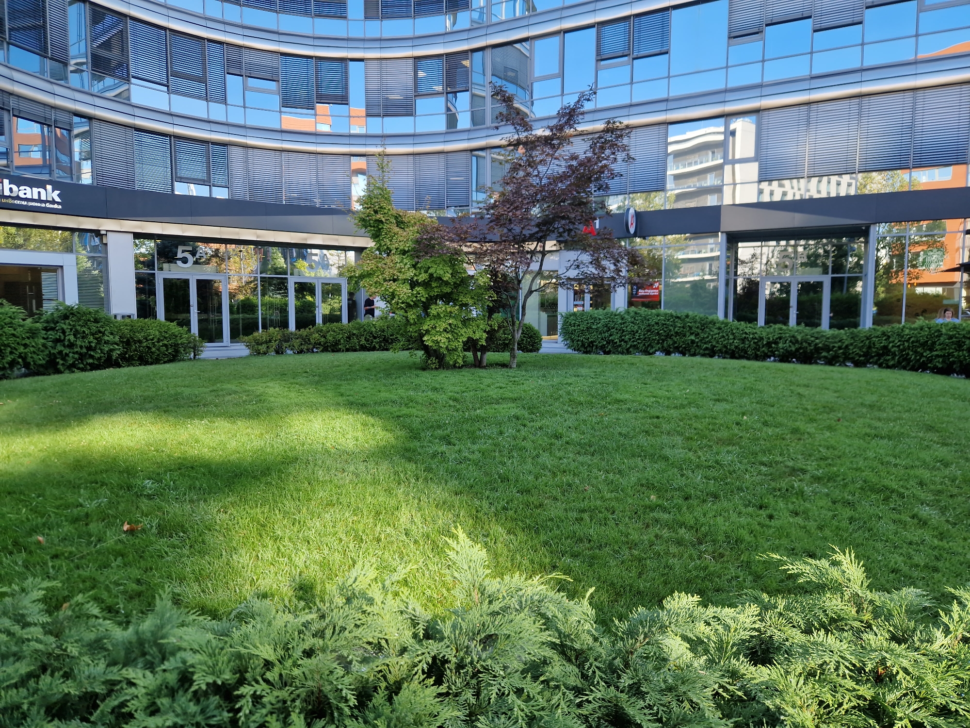 The image shows the exterior of a modern office building in Sofia, Bulgaria, with a well-maintained green lawn in front. The building has a curved facade of glass and metal, with rows of horizontal blinds on the windows. A sign on the building reads "bank". In the foreground, there is a patch of green grass with a small tree in the middle and some bushes on either side. The sun is shining, casting shadows on the lawn. The scene appears to be peaceful and orderly, suggesting a professional business environment.