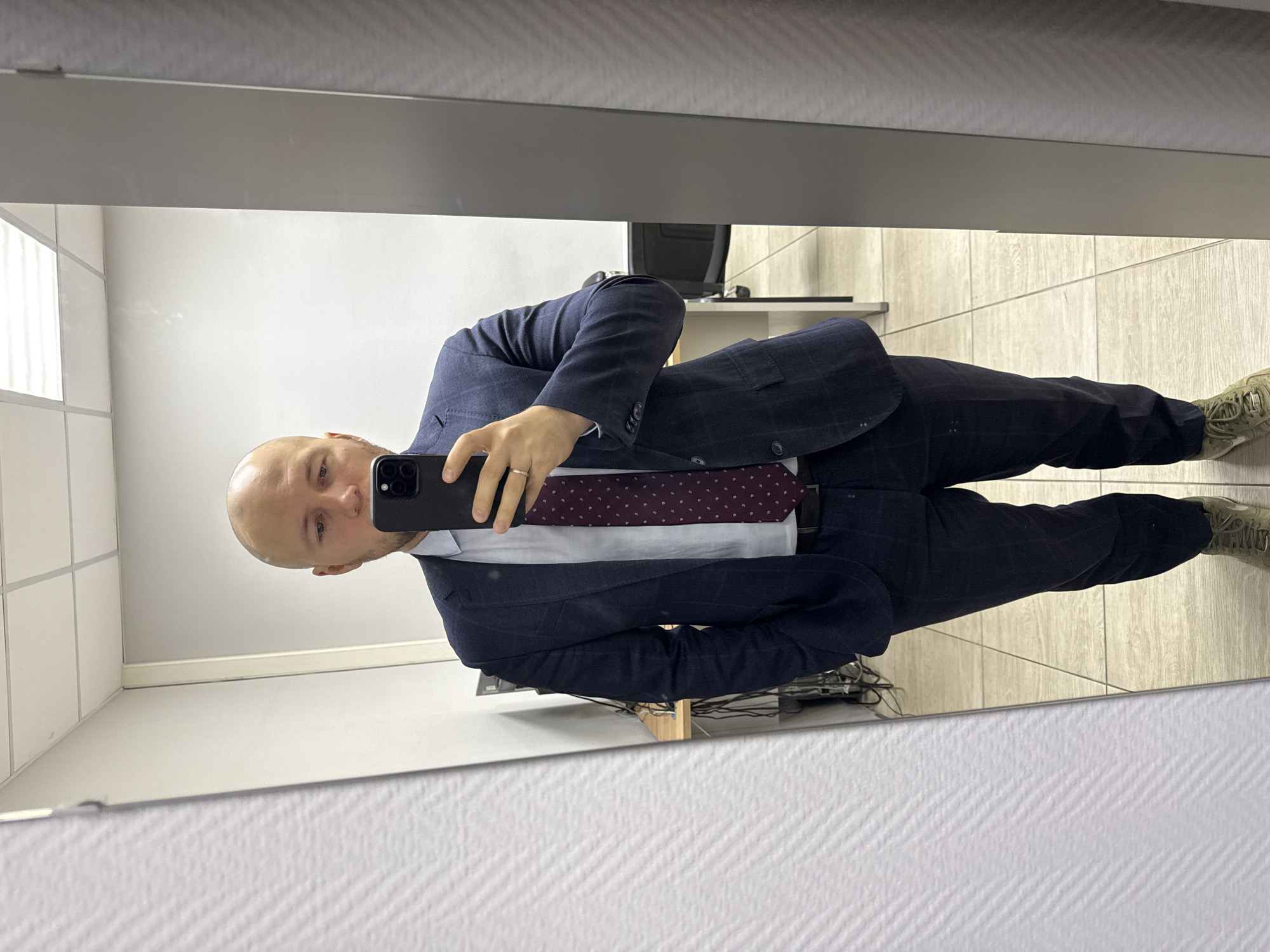 A bald man wearing a blue suit, a white shirt, and a patterned burgundy tie is taking a selfie in a mirror. He is holding a black smartphone in his left hand. His shoes are tan sneakers. The background includes a white wall, a tiled floor, and a partial view of a desk and a computer monitor. The location is likely an office restroom based on the decor and layout. There is no definitive evidence that the location is Krasnoyarsk, Russia.