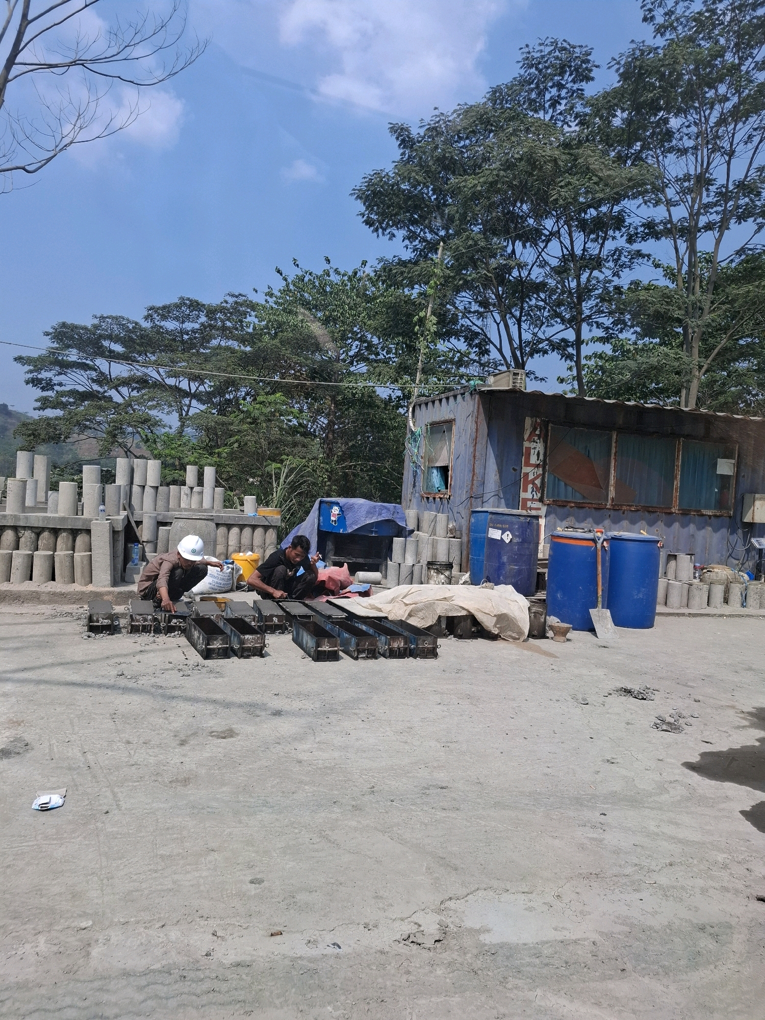 Two men are working at a construction site in Cijangkar, Indonesia. In the foreground, they are kneeling beside a row of metal molds, which appear to be for casting concrete. One man, wearing a white hard hat and a brown shirt, is looking down at the molds, possibly preparing them or checking their alignment. The other man, in a black shirt, is actively engaged with the molds, perhaps placing something inside them. A large bag, likely containing cement or similar material, is placed between them.
Behind the men, a blue makeshift container structure serves as a shelter or storage unit, with windows and a corrugated metal roof. Several large blue barrels are positioned near this structure. In the background, stacks of pre-cast concrete cylinders are neatly arranged. Trees and foliage surround the site, with a clear blue sky overhead. The ground is a mix of concrete and dirt, with some debris scattered around. The overall scene depicts a typical outdoor construction or manufacturing area.