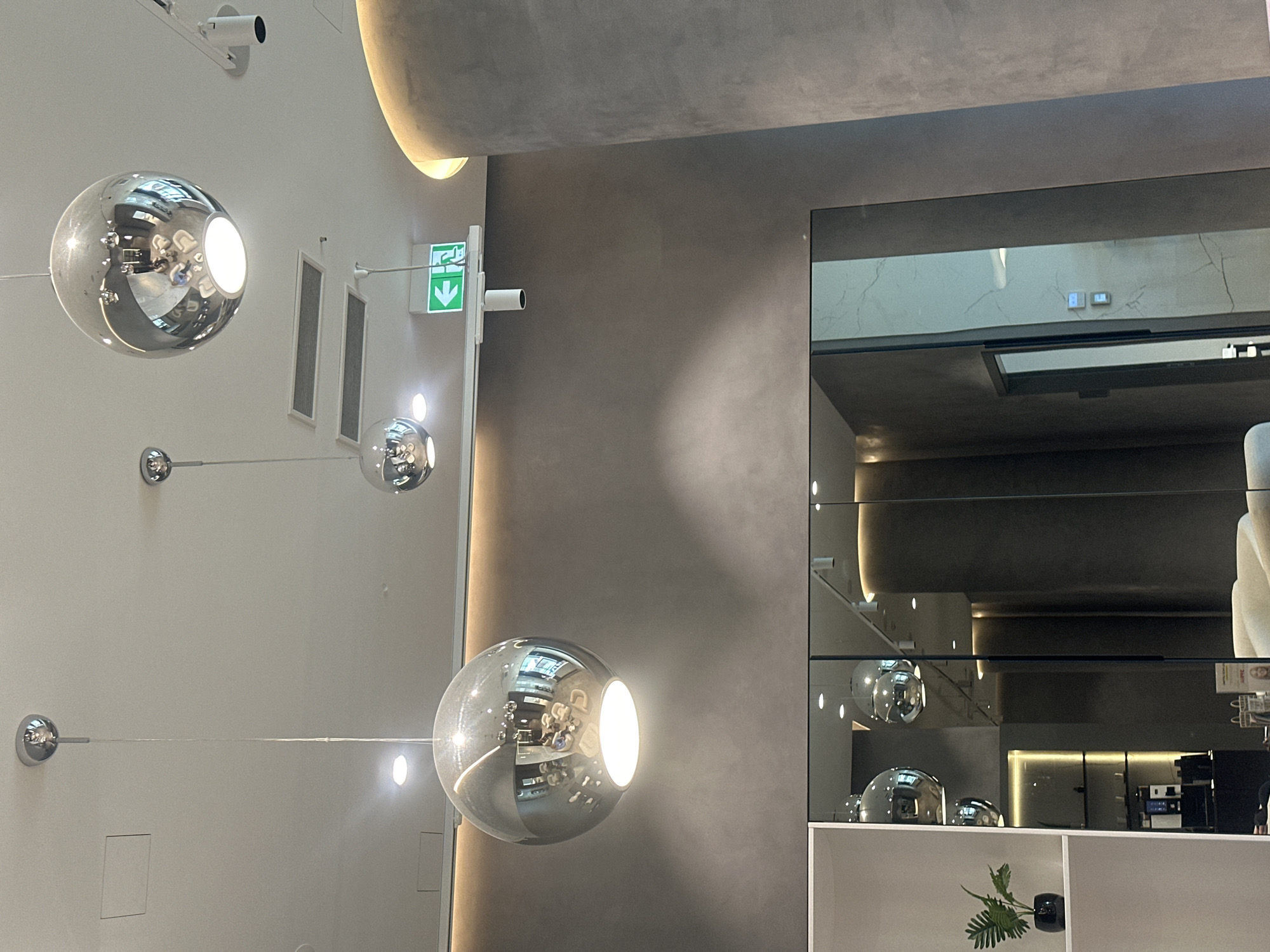 The photograph captures an interior space in Riga, Latvia, featuring modern design elements. There are spherical, reflective light fixtures hanging from a white ceiling, with one prominently in the foreground. An emergency exit sign is visible near the ceiling. Adjacent to the white wall, there is a gray wall or structural component. A mirrored wall reflects the interior, including the hanging light fixtures and elements of what appears to be a café or restaurant setting, with a shelf holding objects and a small plant. No people are directly visible in the scene.