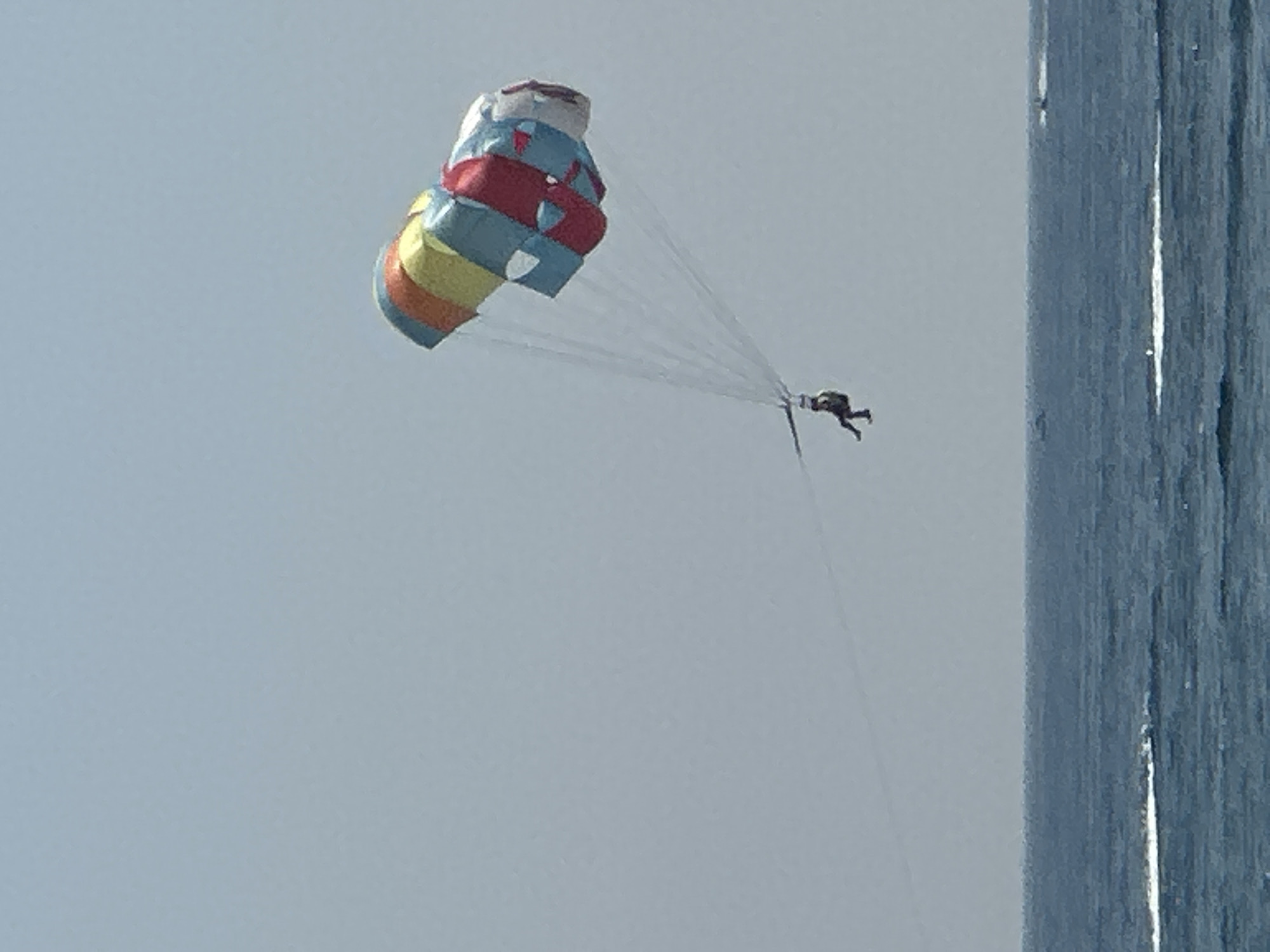 The image captures a bright, clear day with a vast expanse of calm, blue water. In the sky, a person is parasailing, suspended by a colorful parachute. The parachute is a vibrant mix of red, yellow, blue, and white segments. Below the parachute, the parasailer appears to be wearing dark clothing and is seated in a harness, with their legs dangling freely. Thin, white lines, presumably the tow ropes, connect the parasailer to an unseen boat. The horizon is clearly defined, separating the deep blue of the sea from the pale, overcast sky. In the distance, on the water, faint white streaks suggest other boats or watercraft activity, though they are not distinct enough to identify. The overall scene is tranquil and suggestive of recreational activity. The location is the waters off the coast of Boreti, Montenegro.