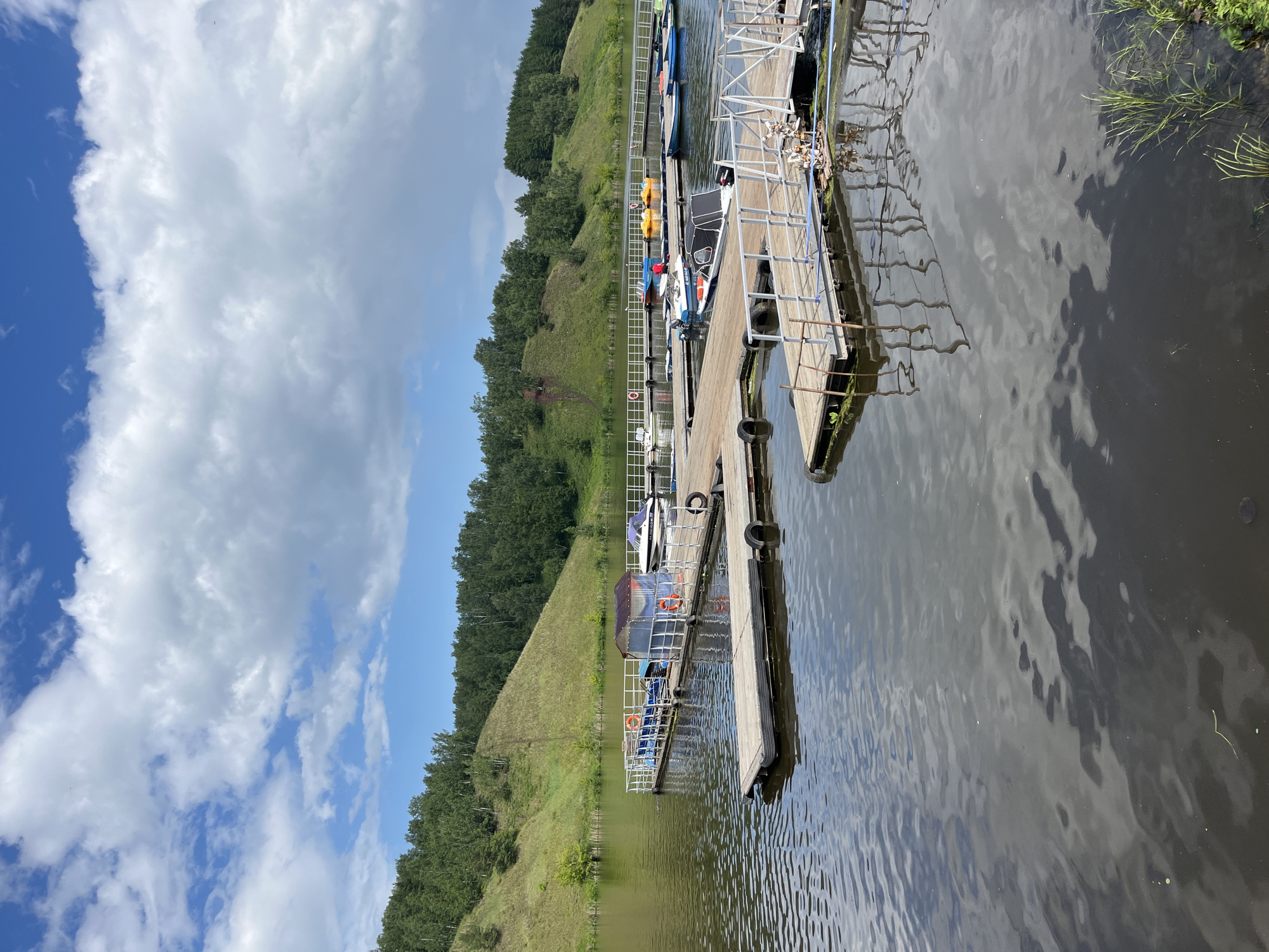 This image depicts a serene lakeside scene at a boat rental station located on the Iset River in Kamensk-Uralsky, Russia. In the foreground, a wooden pier extends into the dark, reflective water, with various small boats and paddle boats docked along its length. The pier is equipped with railings and a small covered structure. In the background, lush green hills covered in trees rise from the opposite bank of the river. The sky above is a mix of blue and white clouds, suggesting fair weather. The overall impression is one of a peaceful recreational area on a summer day.