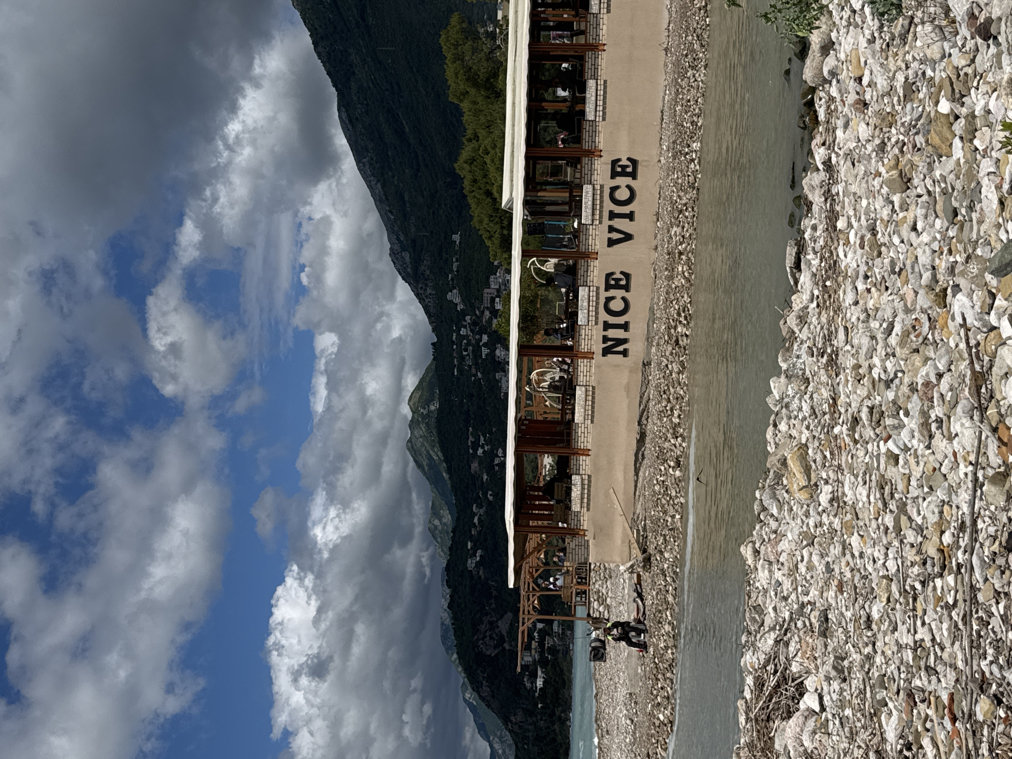 The image shows a scene in Bar, Montenegro, featuring a restaurant called "Nice Vice" built alongside a rocky riverbed. The restaurant has a simple, open structure with multiple levels, each offering seating areas. Patrons can be seen dining and relaxing at tables on these levels. Adjacent to the restaurant is a river with calm water. The riverbank is covered with rocks and pebbles. A mountain range rises in the background, partially obscured by clouds. The sky above displays a mix of blue patches and dense, white clouds. In the foreground, a motorbike is parked near the riverbank. The overall impression is that of a relaxed dining experience in a scenic, natural setting.
