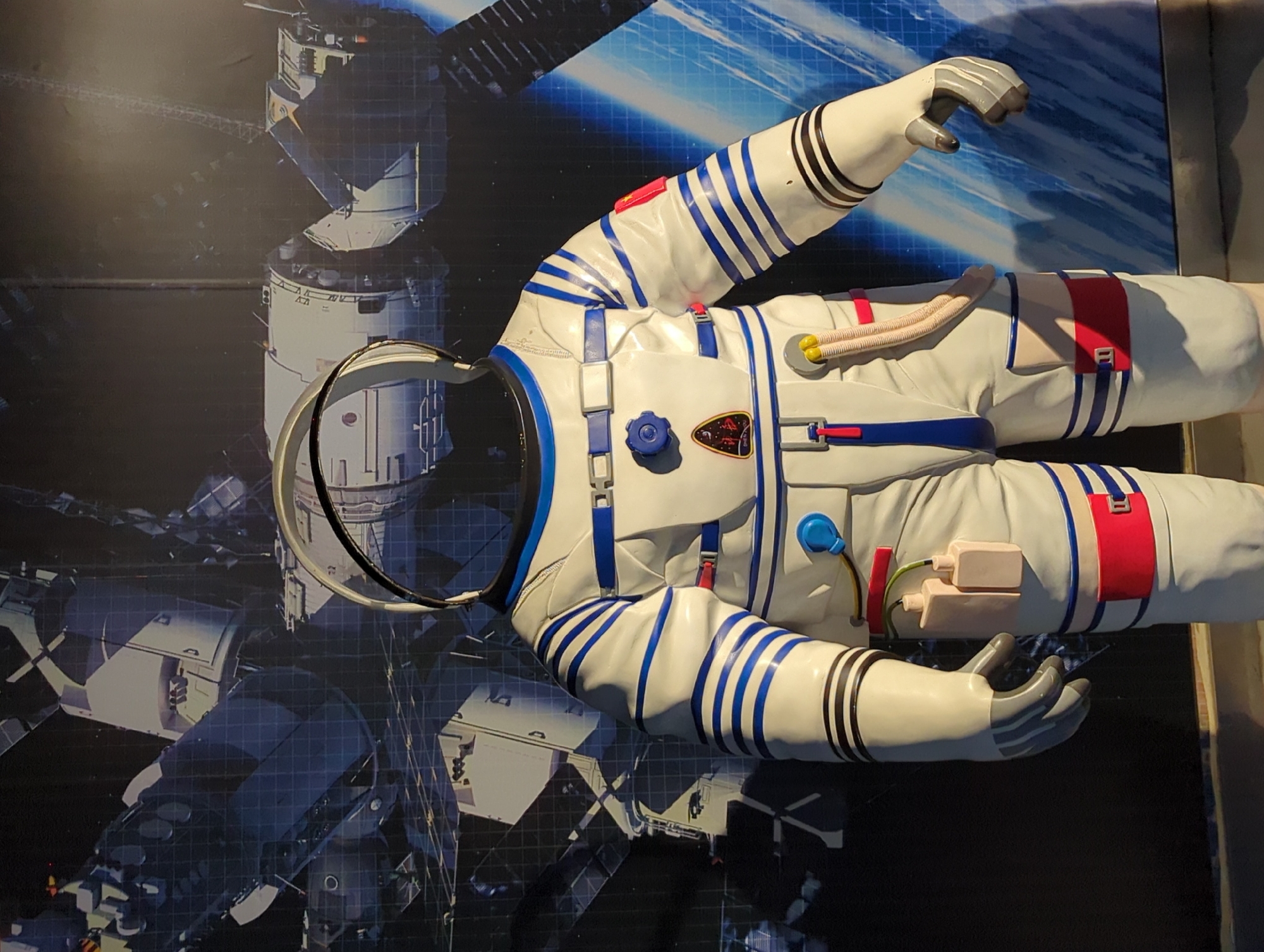 The image displays a full-body mannequin wearing a white and blue astronaut suit, positioned in front of a large, detailed mural. The mural depicts a space station with various modules and a blue grid overlay, suggesting an exhibition or educational display. The astronaut suit features prominent blue stripes on the arms and legs, red accents, and several attached components like tubes and what appear to be small containers. The helmet is transparent, revealing the empty interior of the suit. There are patches on the suit, including one with what looks like a Chinese flag and another emblem with Chinese characters. The setting appears to be indoors, with a dark floor visible at the bottom of the frame. The mural's backdrop suggests a theme related to space exploration. The location is identified as 江北城街道, China, likely within a museum or science center given the nature of the exhibit.