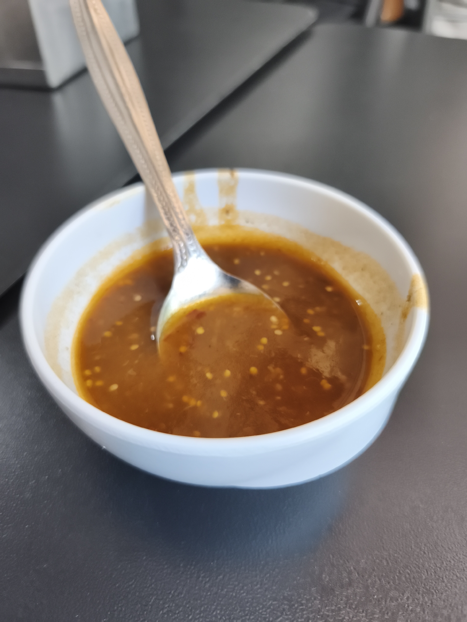 A close-up photograph shows a white bowl filled with a brown sauce. A silver spoon is partially submerged in the sauce. The sauce contains small, light-colored seeds or granules. The bowl is sitting on a dark gray surface. The setting appears to be a table in Mexico City, Mexico. No people are visible.