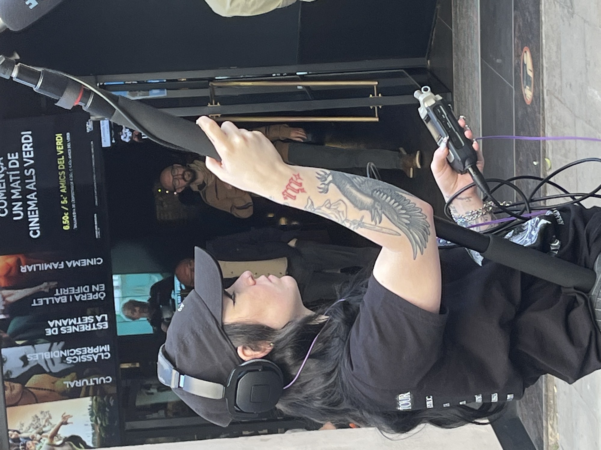 A person is depicted from a side profile, operating sound recording equipment. The individual wears a black baseball cap, black headphones, and a black t-shirt with partially visible white text that includes "TOUR" and "WHIC". Their right arm, extended forward to hold a boom microphone pole, features a prominent tattoo of a large winged creature. Their left hand, adorned with long, light-colored nails and bracelets, holds a portable audio recorder connected by purple and black cables.
The setting appears to be the entrance of a cultural venue or cinema, indicated by large promotional screens or posters in the background. The text on these displays is primarily in Catalan/Spanish, featuring titles and sections such as "CULTURAL," "CLASSICS IMPRESCINDIBLES" (Classic Must-sees), "ESTRENES DE LA SETMANA" (Premieres of the Week), "OPERA I BALLET EN DIFERIT" (Opera and Ballet Delayed Broadcast), and "CINEMA FAMILIAR" (Family Cinema). A notable headline reads "COMENÇA UN MATÍ DE CINEMA ALS VERDI" (A Morning of Cinema at Verdi Begins), with pricing details "6.50€ / 5€ AMICS DEL VERDI" (6.50€ / 5€ Friends of Verdi), suggesting a cinema named "Verdi" in Barcelona, Spain. Other blurred figures are visible inside the entrance. The lighting suggests it is daytime. The overall scene indicates a media production or event coverage at a cultural center or cinema.