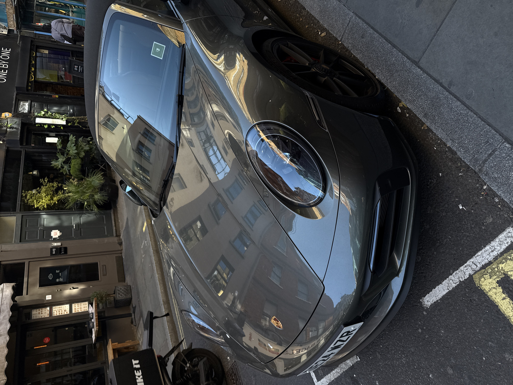 A sleek, dark grey Porsche sports car, likely a 911 convertible, is prominently parked on the side of a street in what appears to be London. The car's front and passenger side are visible, angled towards the viewer. Its highly polished surface reflects the facades of surrounding buildings, showing their windows and architectural details. The bright reflections suggest a clear, sunny daytime.
In the background, a building with a black facade features a sign that reads "ONE BY ONE." Further down the street, another building with large windows is reflected in the car's windshield and hood. A person wearing a light-colored shirt and a backpack is partially visible in the reflection, seemingly walking past.
The street features a grey pavement and asphalt road with distinctive yellow and white lines, indicative of UK road markings. The car's front license plate is partially visible, displaying "24 VZR," confirming the UK location. The Porsche emblem is clearly visible on the car's front hood. On the far left, part of a bicycle or scooter is visible with the text "BIKE IT". A small green sticker with illegible text is seen on the car's windshield. The scene captures an urban street environment with a luxury vehicle.