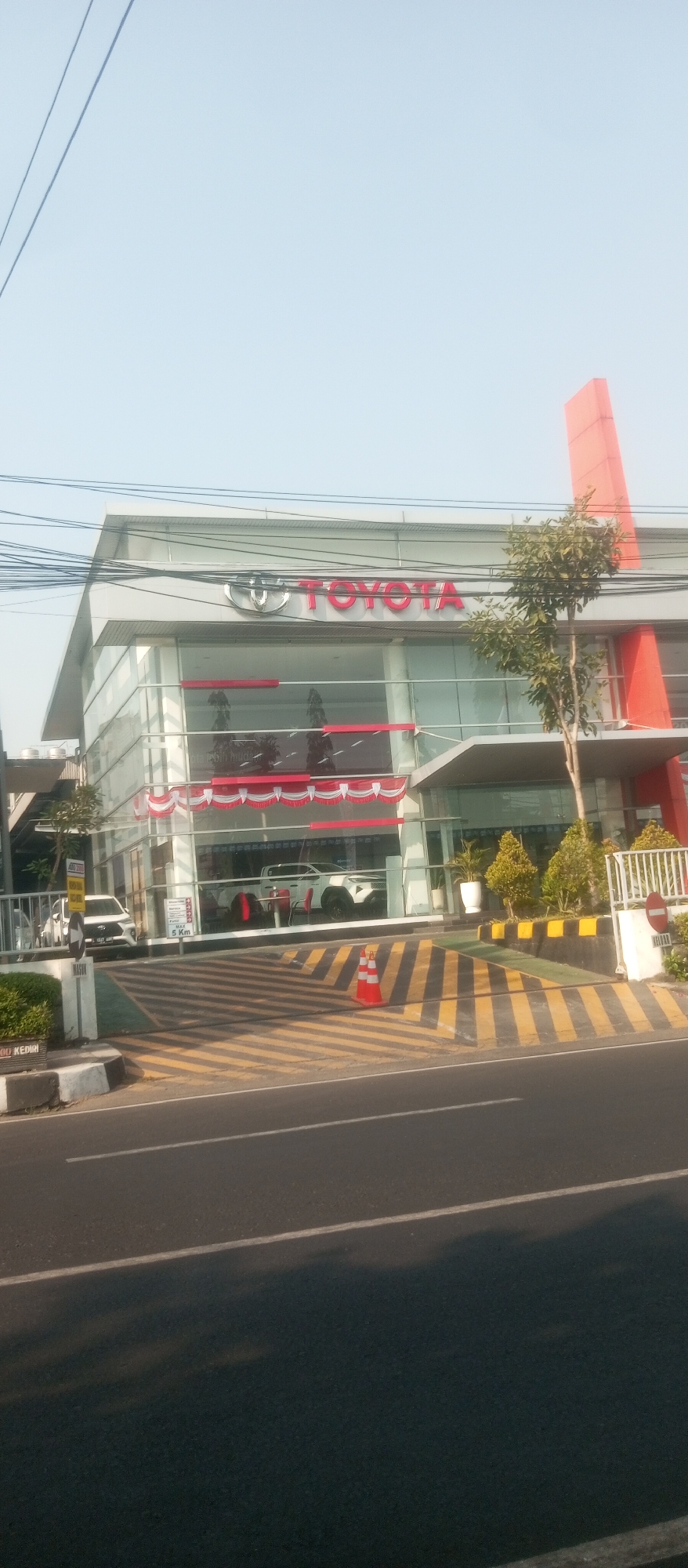 A bright, clear daytime shot captures the exterior of a modern Toyota dealership in Kediri City, Indonesia. The building features a prominent glass facade with a bright red Toyota logo and lettering mounted above. A tall, orange, angular structure rises from the right side of the building, reaching towards the sky. In front of the dealership, a white car is displayed behind the glass. A ramp with diagonal yellow and black stripes leads up to the entrance. A single orange traffic cone rests on the ramp. Several power lines are visible in the sky above the building. The scene is captured from across a road with a white line marking the lane.