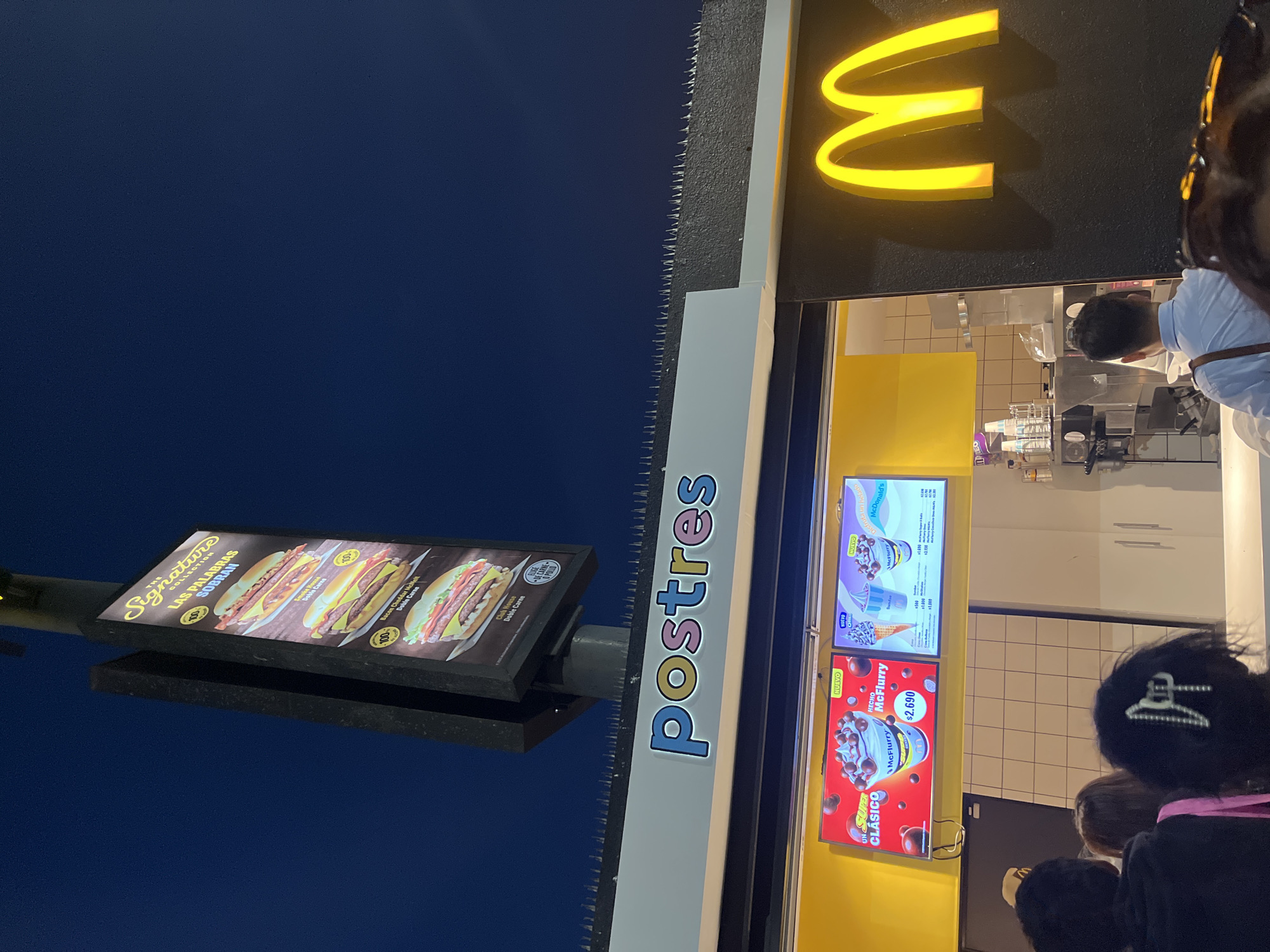 The image captures a McDonald's in Arica, Chile, at night. The restaurant is recognizable by its illuminated golden arches logo. A menu board advertising desserts, specifically McFlurries, is visible near the drive-thru window, along with a sign indicating that the "Postres" (desserts) are being sold. The "Signature Collection" menu board shows images of various burgers. Customers are waiting at the window, and an employee is visible inside. The sky is a deep blue, suggesting it is late in the evening or early morning. The McDonald's building has spikes installed along its roof to deter birds.