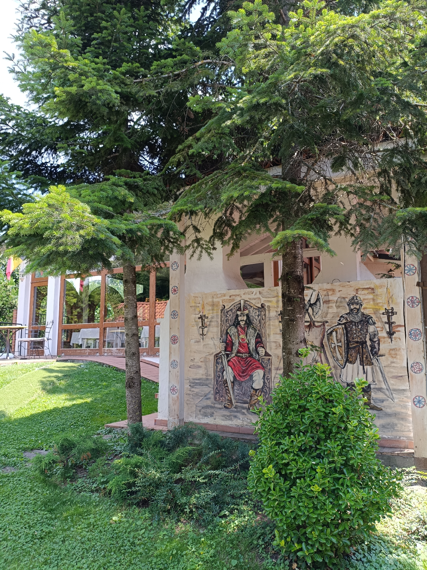 The image shows an outdoor scene in Comana, Romania. In the foreground, a lush green lawn is dotted with clover, leading to a bushy green shrub. A large tree with dark bark and green needles stands in the middle of the frame. Behind the shrub and tree, a white building with a curved archway is visible, featuring a mural painted on its wall. The mural depicts two figures: one man seated on a throne wearing a red robe and crown, and another man standing in armor, holding a shield and sword. Decorative circular patterns are also painted on the building's exterior. To the left of the building, a glass-enclosed area with tables and chairs can be seen, suggesting an outdoor dining space. The background consists of more greenery, including trees and foliage, under a bright sky.