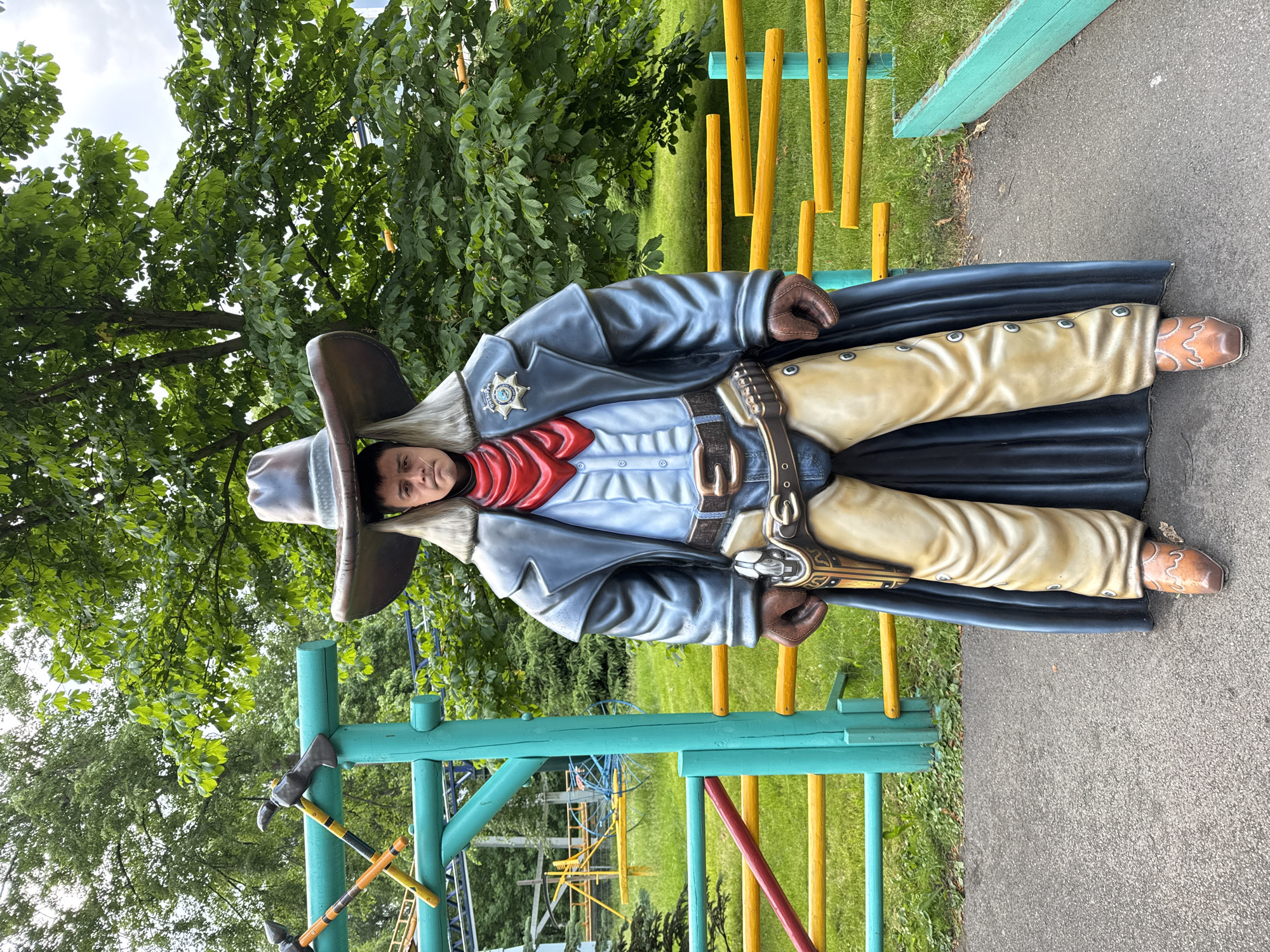 An outdoor, eye-level, medium shot captures a life-sized statue of a cowboy in the center, posing stoically. The statue, dressed in a wide-brimmed hat, red bandana, blue shirt, and a dark coat, features a sheriff's badge on its chest. It holds a pistol in a holster on its right hip, with a bullet belt across its waist. The cowboy statue stands on a paved path, with a grassy area and colorful barriers to its right. In the background, a lush green tree canopy dominates the scene, with the structure of a roller coaster visible to the left. The setting is an amusement park, possibly located in Saint Petersburg, Russia, given the visual cues of similar attractions found in the region.