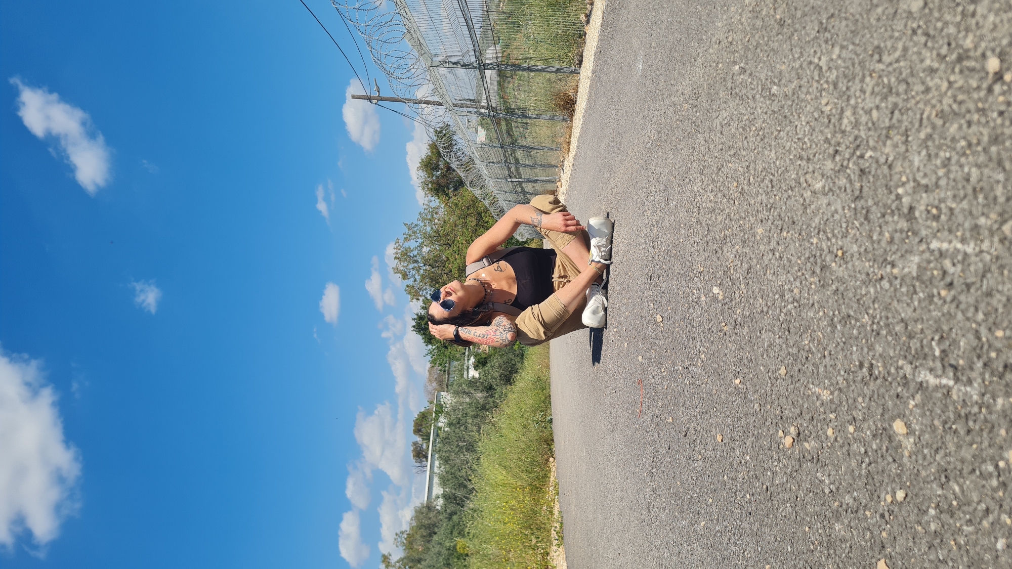 A person wearing a black tank top, tan pants, and white sneakers sits on a gray asphalt road. They are also wearing sunglasses and appear to have tattoos on their arms. The person is positioned between a blue sky with scattered clouds and a fence topped with barbed wire that runs parallel to the road. Vegetation grows along the roadside. The scene appears to be located in the Lachish Regional Council, Israel.