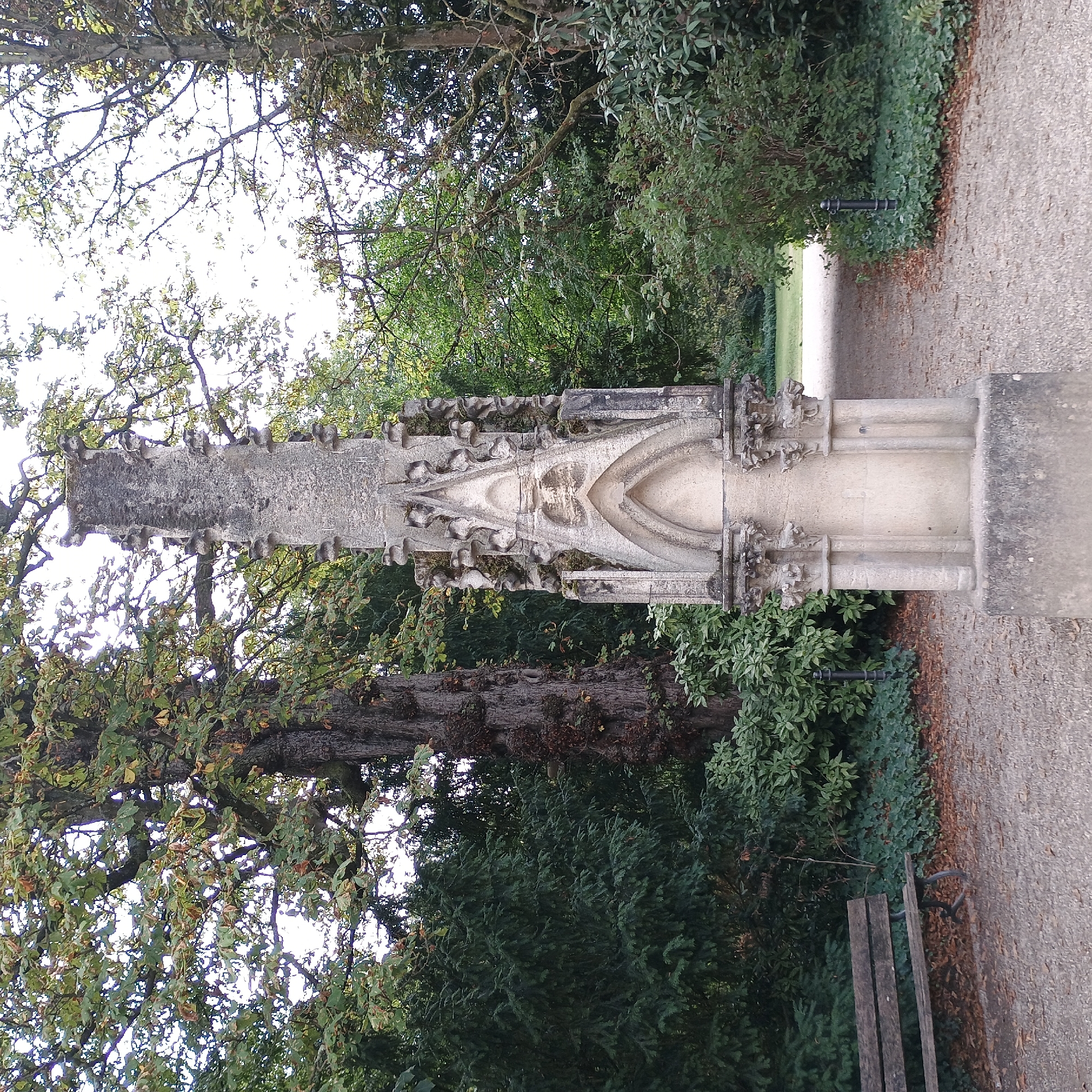A stone monument, weathered and showing signs of moss, stands in the center of the frame. The monument is tall and slender, with Gothic architectural elements, including pointed arches and ornate carvings, suggesting it might be a cenotaph or a decorative garden feature. It is positioned on a gravel path, flanked by lush green trees and shrubbery. To the left of the monument, a wooden park bench is visible, showing wear and tear. The trees behind the monument are a mix of deciduous and evergreen, with some autumn leaves scattered on the ground. The sky is overcast and bright, suggesting a daytime scene. The overall setting appears to be a park or garden, possibly within a historical or landscaped area. This monument is located in the "Kölner Grüngürtel" (Cologne Green Belt), a large park surrounding the city center of Cologne, Germany.