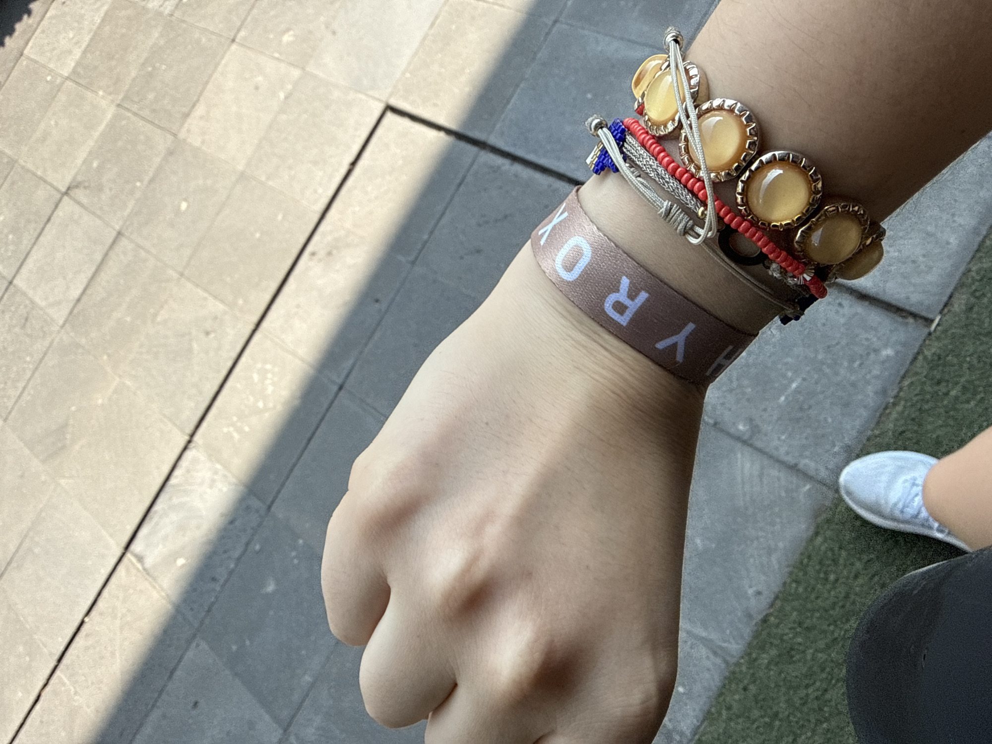 A close-up shot captures a person's left hand and wrist adorned with several bracelets. The wrist features a brown wristband with the text "HYROX" in white letters. Above it, a stack of bracelets includes a silver beaded bracelet, a bracelet with red and blue beads, and a more elaborate bracelet with large, round, yellow-gold stones. The person is wearing a white sneaker and black athletic wear.
The setting appears to be outdoors on a grey tiled surface with a distinct light and shadow pattern. The location is unknown; no detail confirms it is in Mexico City, Mexico.