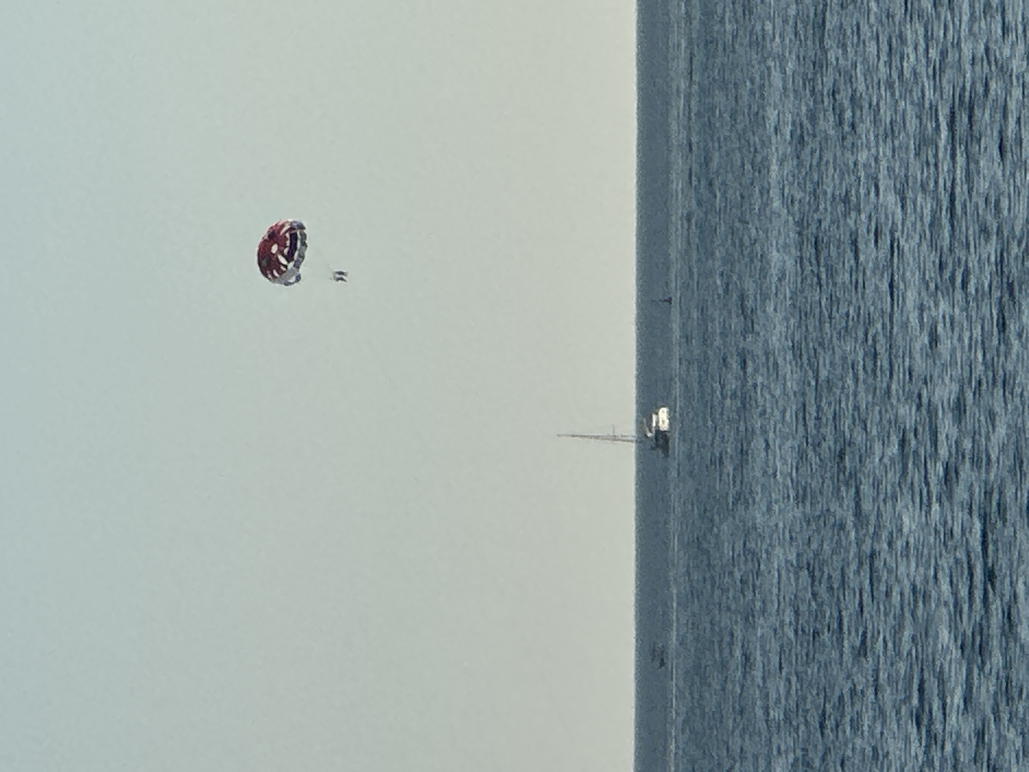The image depicts a wide expanse of blue ocean under a pale, overcast sky. In the upper portion of the frame, a red and white parachute is visible, with two small figures suspended beneath it, engaged in parasailing. Further down, on the horizon, a sailboat with a single mast is white against the blue water. Another smaller boat is also visible in the distance. The image appears to be taken from the coast or a vessel, looking out over the Adriatic Sea, possibly near Budva, Montenegro, known for its coastal activities. The overall scene conveys a sense of leisure and outdoor recreation.