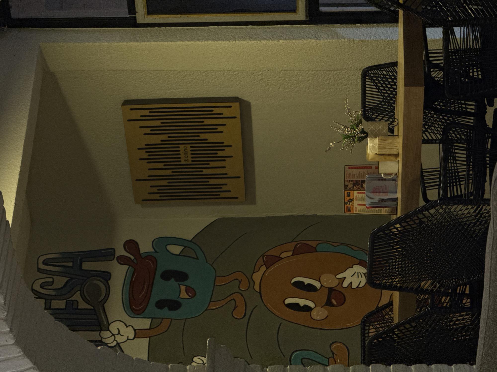 The image depicts an interior wall in San Andrés Cholula, Mexico, featuring a mural with cartoon-like characters: a blue, anthropomorphic coffee mug and an orange, anthropomorphic pancake. A sign with horizontal lines sits above the mural. Stacked black woven chairs stand to the right, partially obscuring the mural and obscuring objects, including a small floral arrangement and menus. The scene is lit artificially.