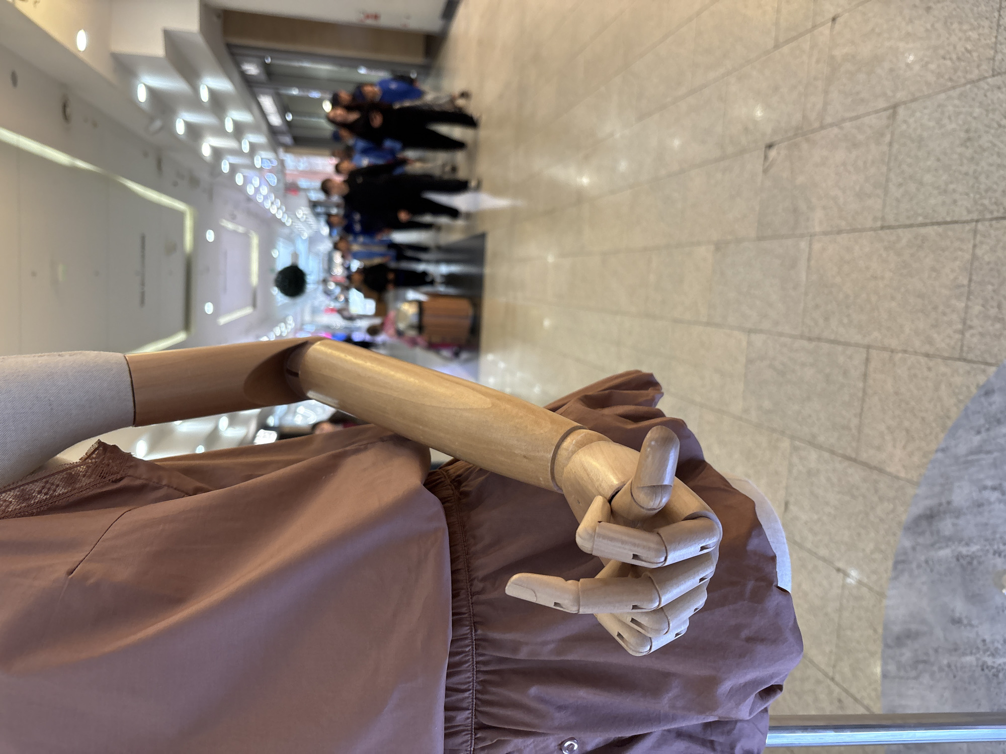 A mannequin with a wooden arm and hand wearing a brown dress is in the foreground. In the background, there is a group of people walking down a corridor with bright overhead lighting. The floor is made of light-colored tiles. The location is inside a building, likely a shopping mall or convention center, potentially in Krasnoyarsk, Russia.