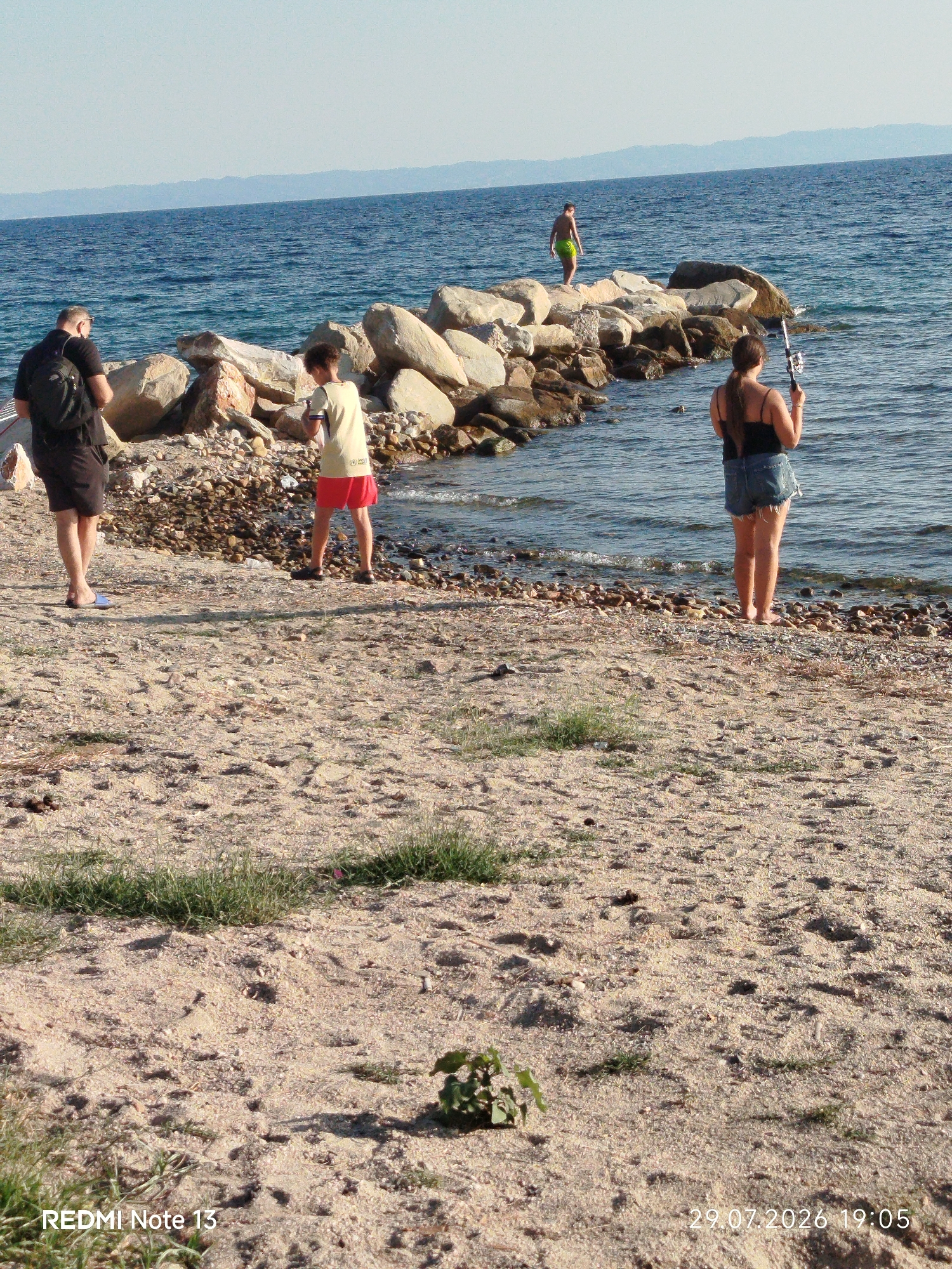 This media file depicts a sunny day at the beach in Sithonia, Greece. The scene is set along the coastline, with a clear blue sky and calm, blue water. A man in a black t-shirt and dark shorts stands on the sandy shore, facing away from the camera, with a black backpack on. Nearby, a young person wearing a light-colored t-shirt and red shorts stands on the rocky edge of the water. Further out on the rocky jetty, another person, a woman with dark hair, is holding a fishing rod and appears to be fishing. The jetty is made of large, light-colored rocks. In the background, a distant shoreline with hills is visible. The overall atmosphere is relaxed and leisurely, suggesting a typical summer day at the seaside. Visible text in the lower left corner reads "REDMI Note 13", and in the lower right corner, "29.07.2026 19:05".