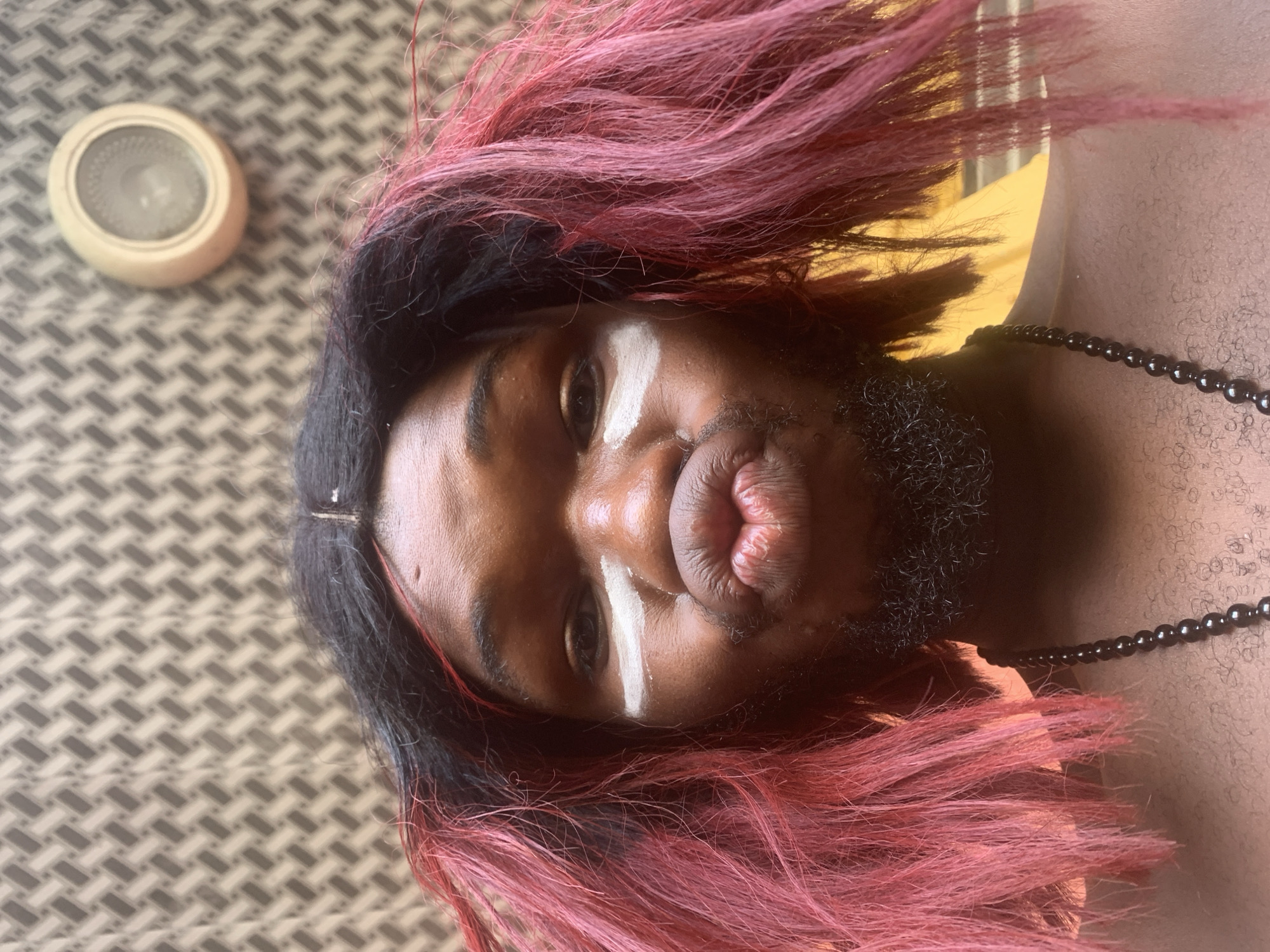 This vertical portrait depicts a person with dark skin and dark hair prominently highlighted with pink, styled with some strands framing the face. The individual also has a short, dark beard. White paint is applied to the face, forming a distinct stripe across the forehead/brow bridge and another mark below the left eye. The person is looking directly at the camera with their lips pursed in a "duck face" or kissy face expression. They are wearing a simple black beaded necklace around their neck.
The background features a light-colored surface with a repeating geometric pattern of grey and white, resembling a woven mat or textured wall. A blurred yellow-orange area is visible on the right side, suggesting another surface or wall. In the upper left, a circular, off-white object with an intricate patterned center, possibly a thermostat or a small speaker, is partially visible. The lighting is bright and natural, indicating a daytime setting indoors in Nkpor, Nigeria. No text is visible in the image.