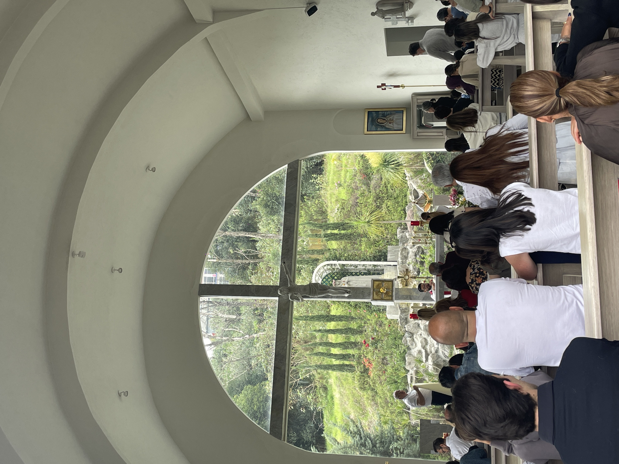 The image depicts the interior of a chapel in Huixquilucan de Degollado, Mexico. Several rows of people are kneeling or sitting in pews facing forward. A large, arched window reveals an outdoor religious scene with a prominent crucifix and a statue, surrounded by greenery and foliage. Inside the chapel, religious artwork hangs on the wall. The ceiling features a series of concentric arches.