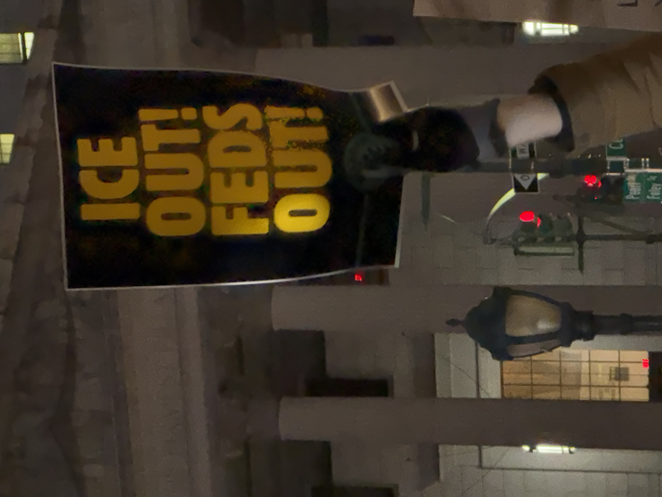 This nighttime image captures a protest or demonstration taking place on a city street, likely in New York City. A person, whose torso and arm are visible on the left side of the frame, holds up a black protest sign. The sign features bold yellow text that reads, "ICE OUT! FEDS OUT!". The person appears to be wearing a dark jacket or hoodie with a lighter-colored sleeve.
The background depicts an urban setting at night. Several buildings are visible, some with illuminated windows, suggesting a dense city environment. A dark, ornate streetlight stands on the right, providing some illumination. Multiple traffic lights are visible in the distance, all displaying red lights, indicating either a stop or an intersection. Partially legible street signs in the upper right hint at "Pearl" and "Foley," which are specific street names found in Lower Manhattan, New York City, near Foley Square, an area frequently associated with protests due to its proximity to government and court buildings. The overall ambiance is dark, with artificial lights cutting through the night. The visible text on the sign expresses a clear message of opposition to Immigration and Customs Enforcement (ICE) and federal agencies.