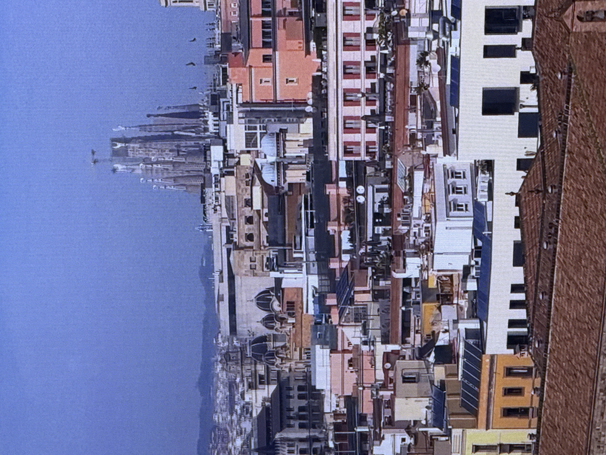 The image depicts a panoramic view of a densely built urban landscape, characterized by a multitude of buildings with varying architectural styles and colors. In the background, a distinctive, unfinished Gothic-style cathedral with tall spires dominates the skyline, suggesting it might be the Sagrada Familia in Barcelona, Spain. The middle ground features a cluster of buildings, some with domed roofs and ornate facades, while others are more modern, with flat roofs and multiple windows. The foreground is filled with a mix of residential and commercial structures, many with terracotta tile roofs. Solar panels are visible on some of the rooftops, indicating a blend of traditional and contemporary infrastructure. The sky is a clear, pale blue, typical of a bright, sunny day. The overall scene conveys a sense of a bustling, historic city with a mix of old and new architecture. The image does not depict the city of Dubai, United Arab Emirates.